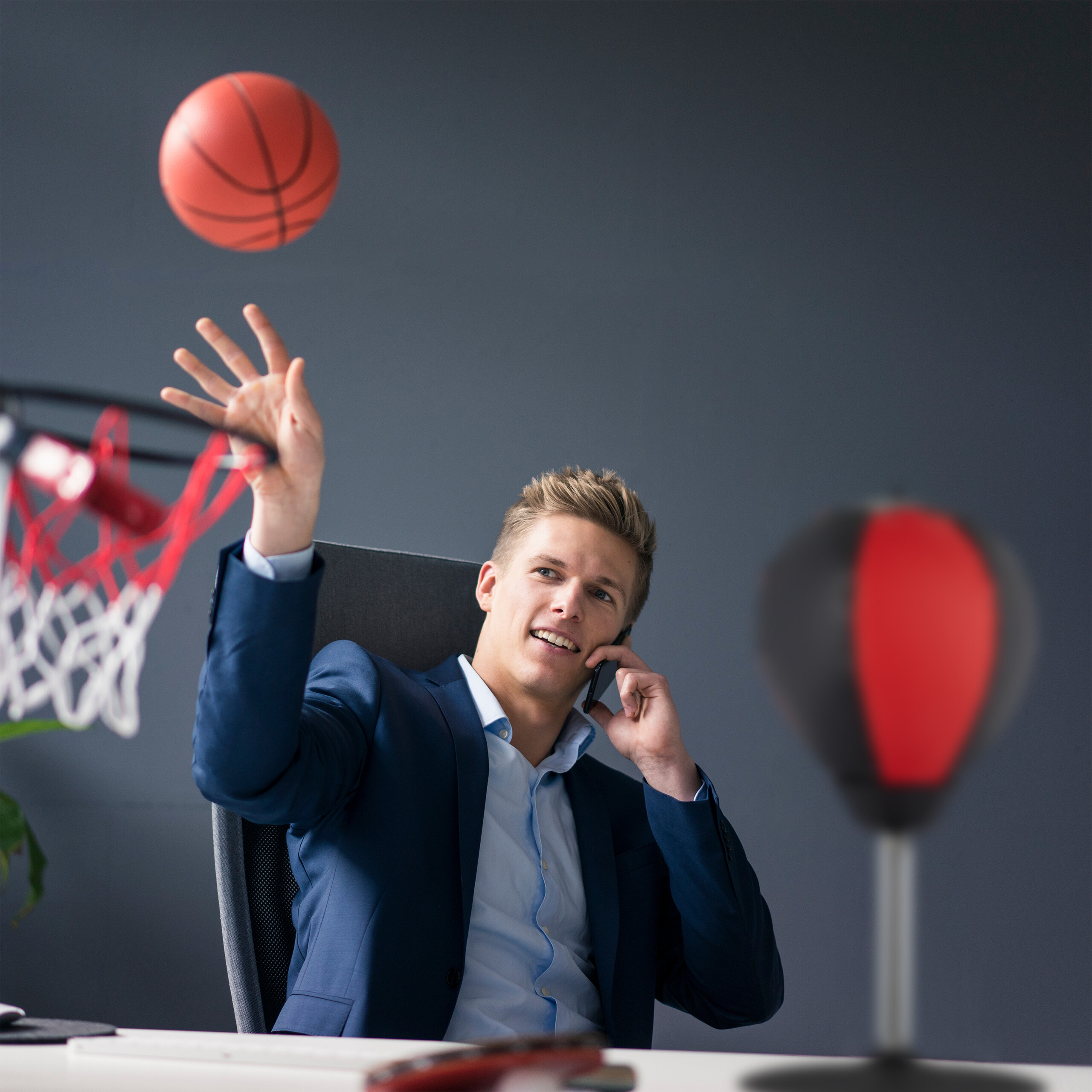 Ein Geschäftsmann im Anzug am Schreibtisch telefoniert und wirft einen Basketball in den Korb.