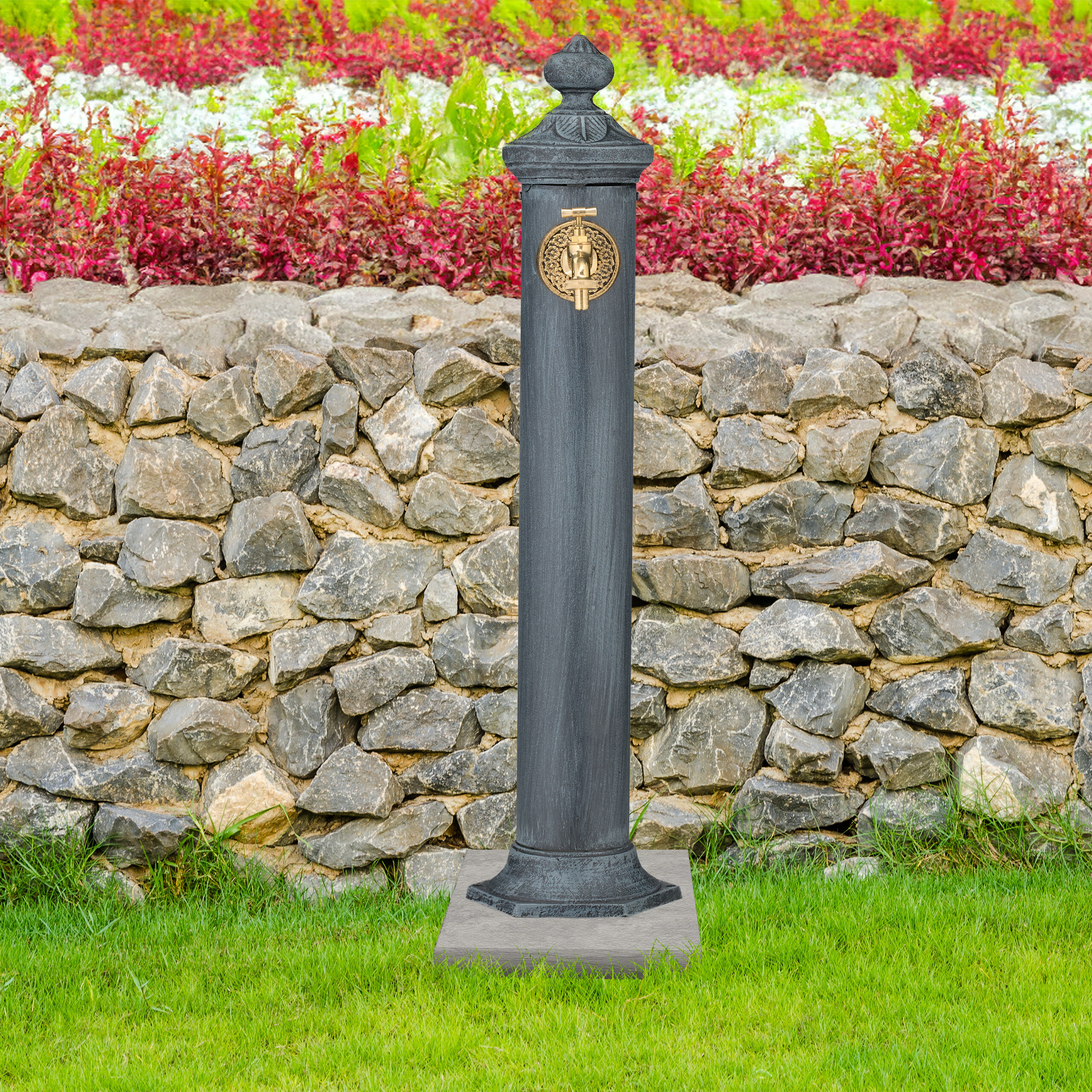 Ein grauer Gartenbrunnen mit goldenem Wasserhahn vor einer Steinmauer und Blumen.