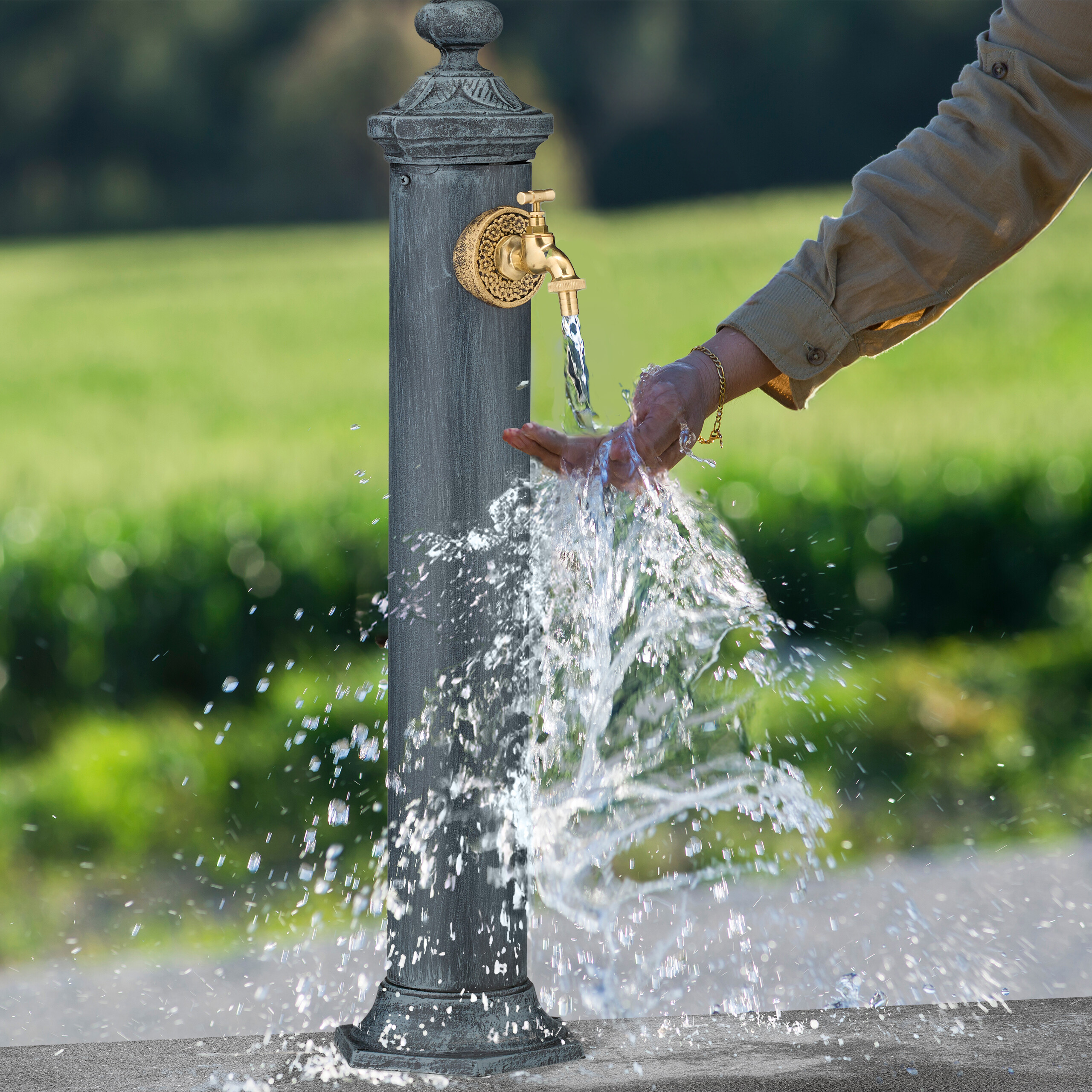 Person wäscht Hände unter fließendem Wasser an einem grauen Gartenbrunnen mit goldenem Hahn.