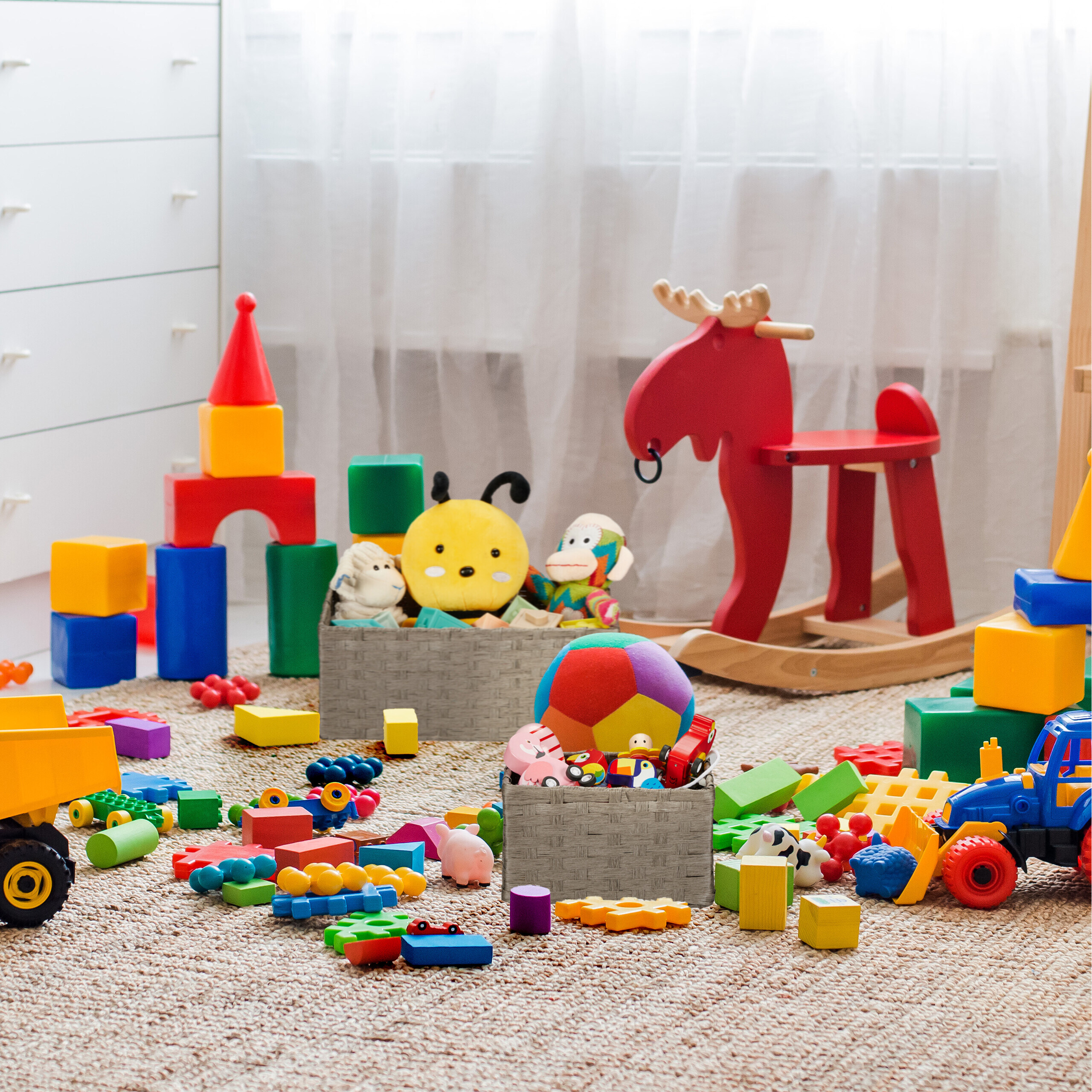 Bunte Spielsachen, Bausteine, ein gelbes Insektenkissen und ein rotes Schaukelpferd liegen auf dem Teppich.