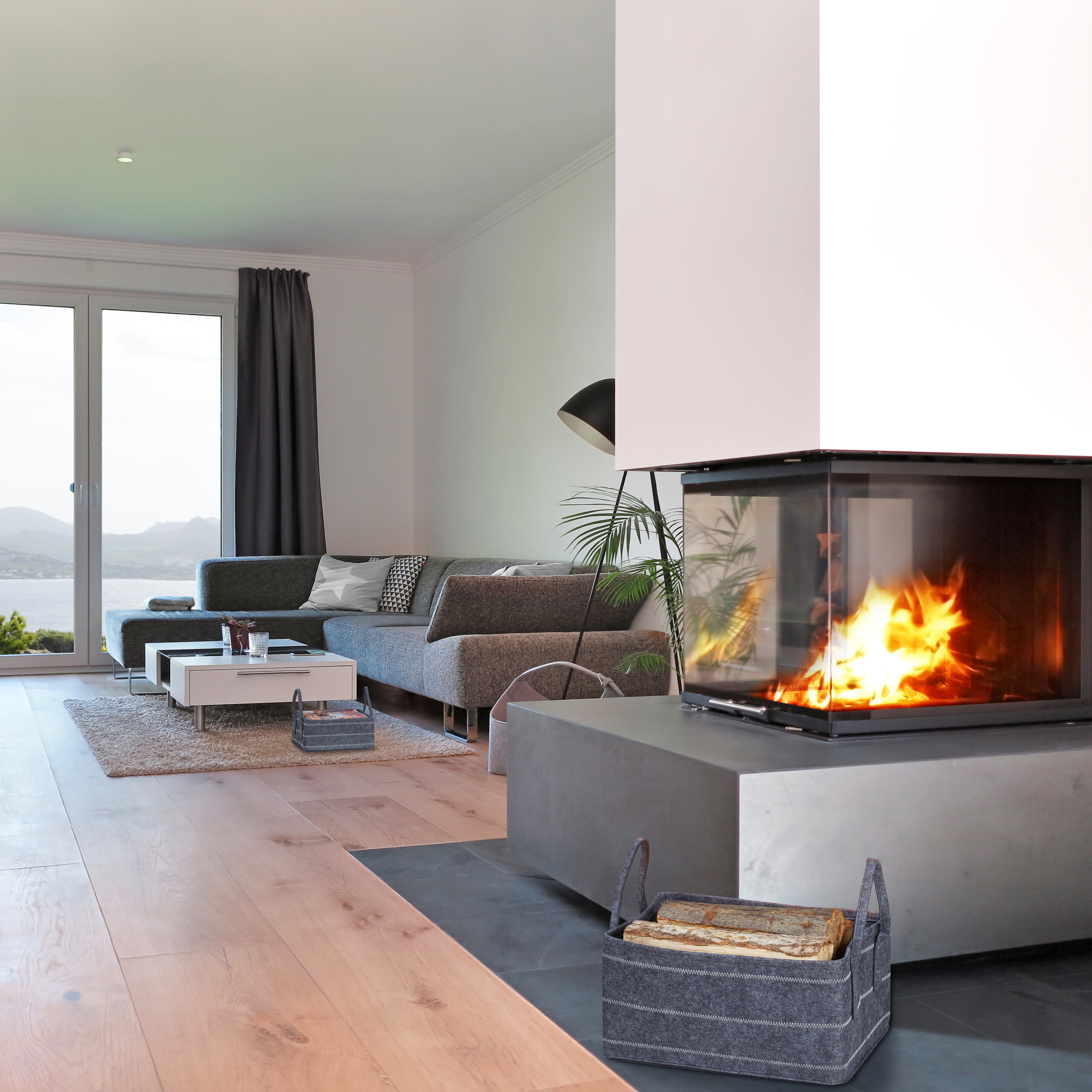 Ein modernes Wohnzimmer mit einem gläsernen Kamin, hellem Feuer, grauem Sofa und Seeblick durch Glastüren.