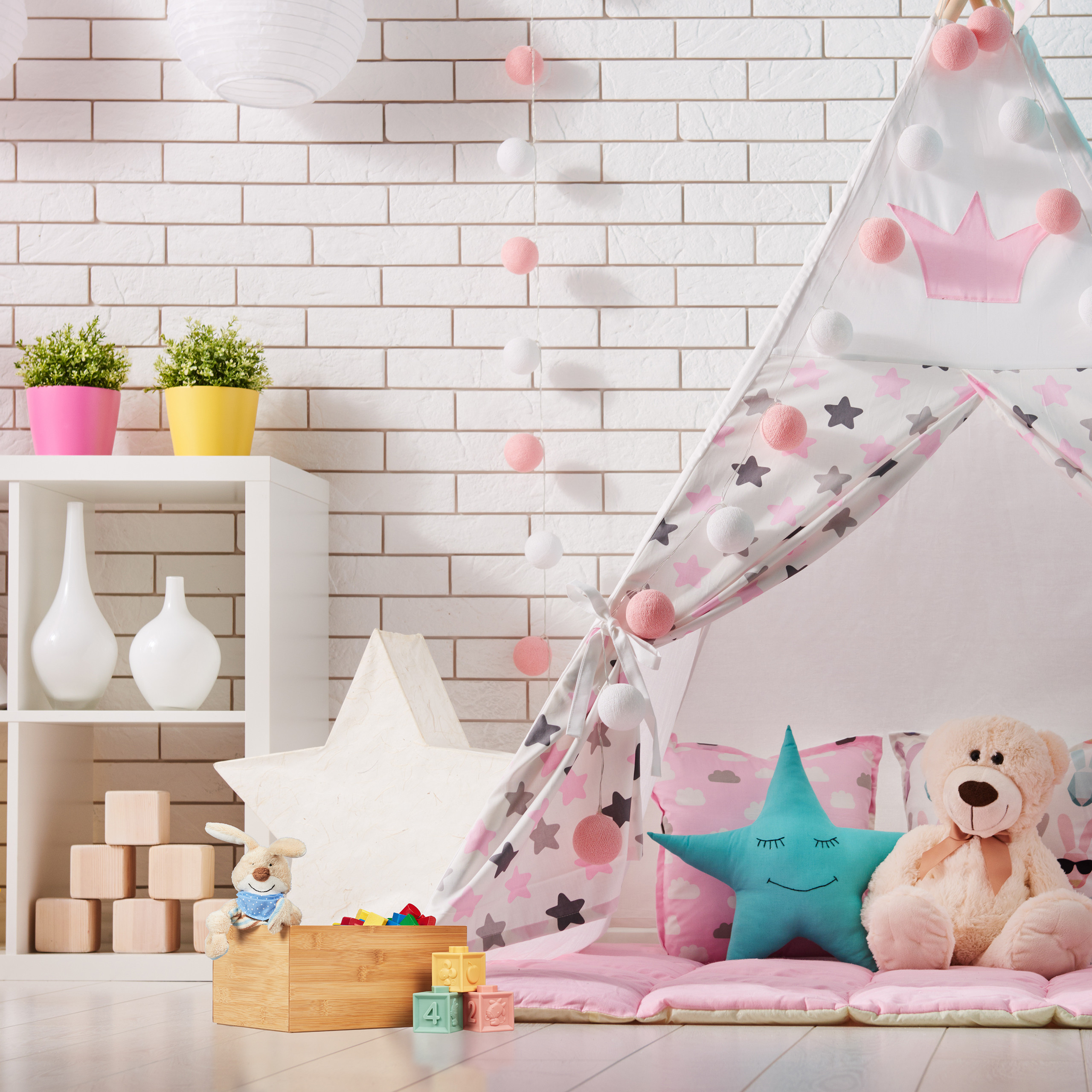 Ein Kinderzimmer mit weißem Spielzelt, einem Teddybär, Kissen und einem Regal mit Pflanzen an einer Ziegelwand.