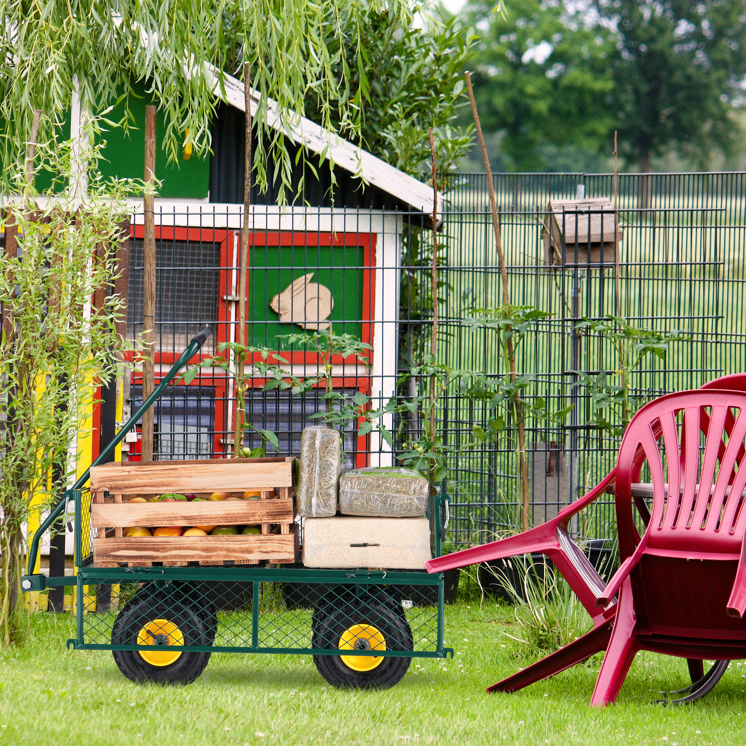 Ein grüner Gartenwagen mit Holzkiste voller Obst und Heuballen im Gras, daneben rote Plastikstühle.