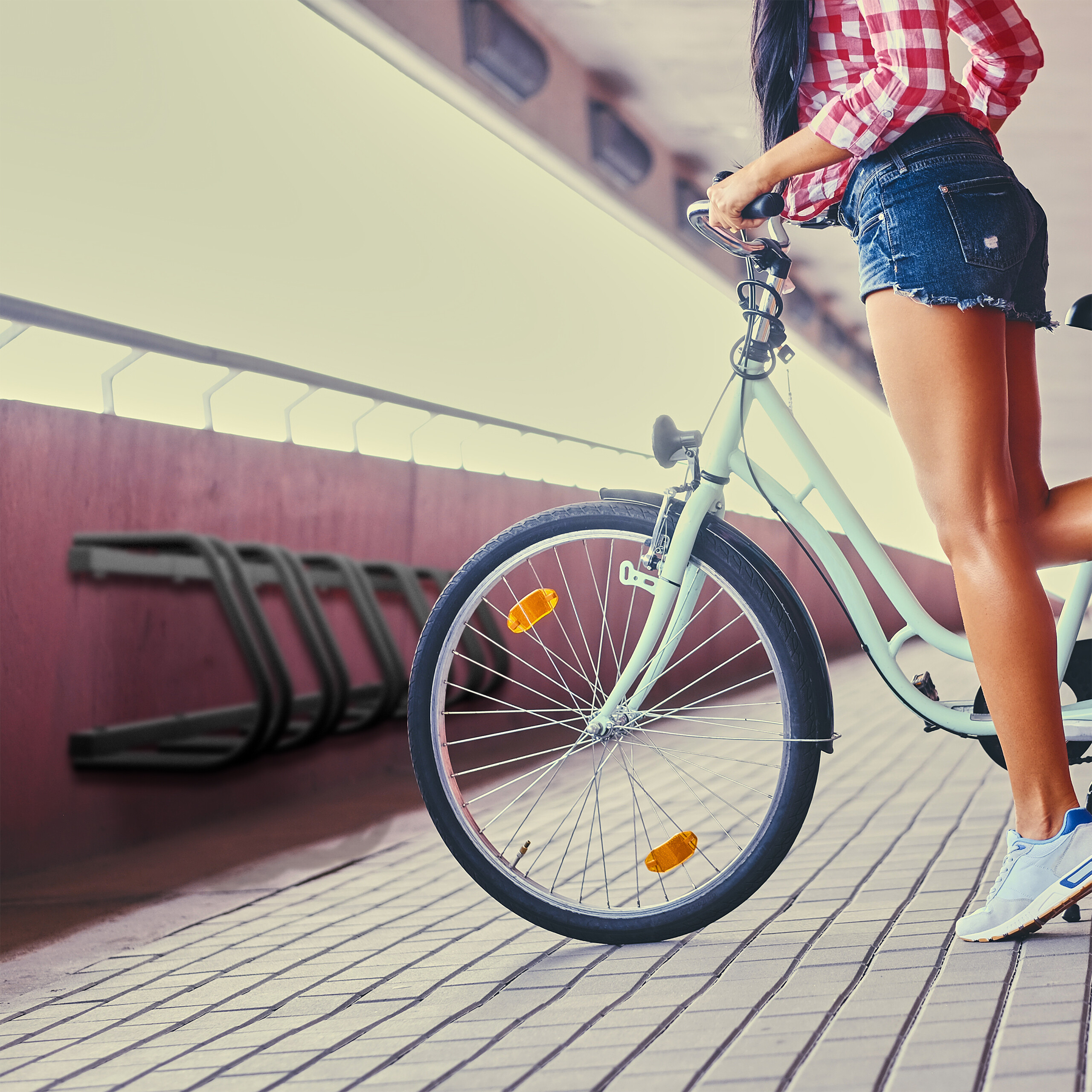 Eine Frau in rotem Karohemd und Jeansshorts steht mit einem hellblauen Fahrrad auf einer gepflasterten Brücke.