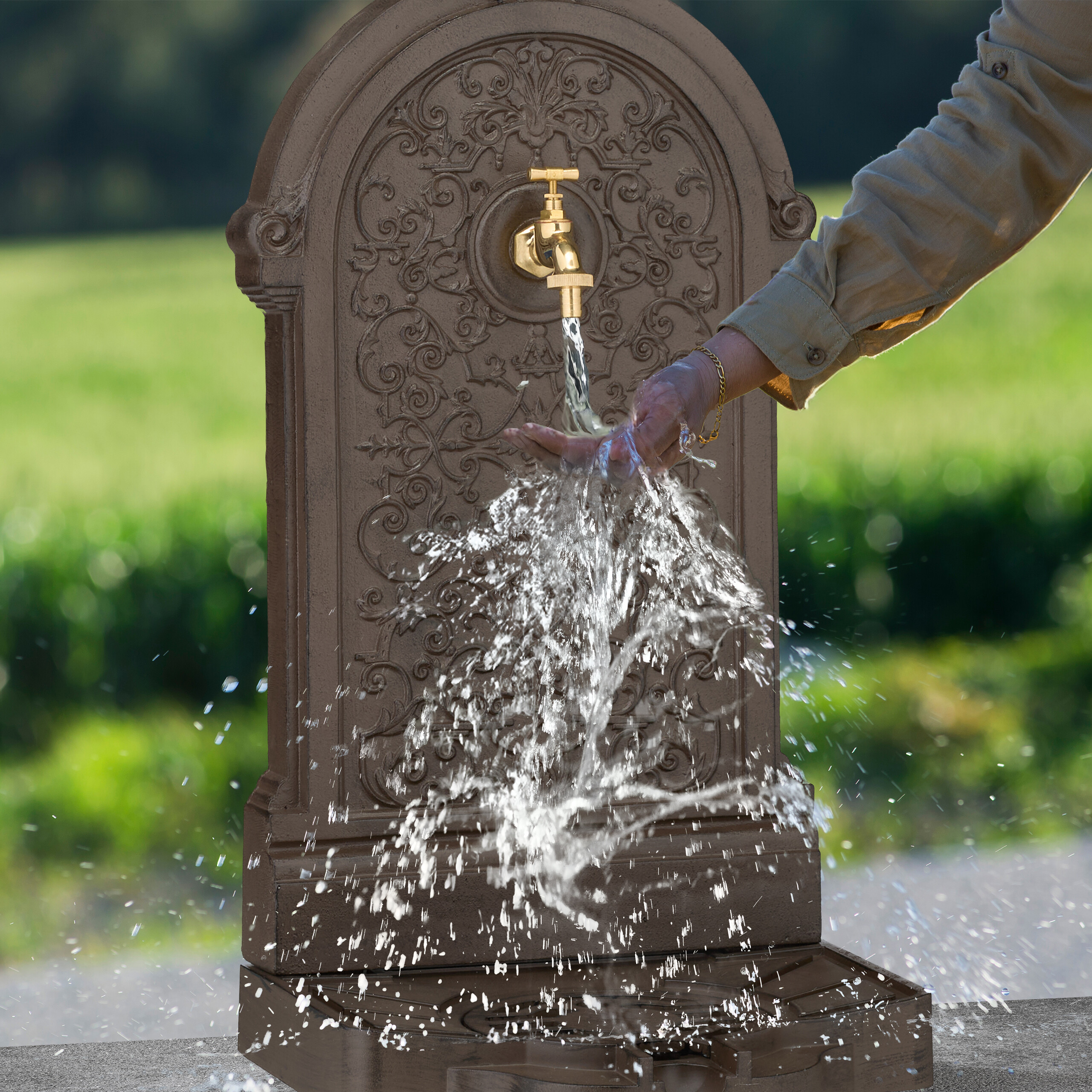 Eine Hand wäscht sich unter dem fließenden Wasser eines verzierten, braunen Wandbrunnens im Freien.