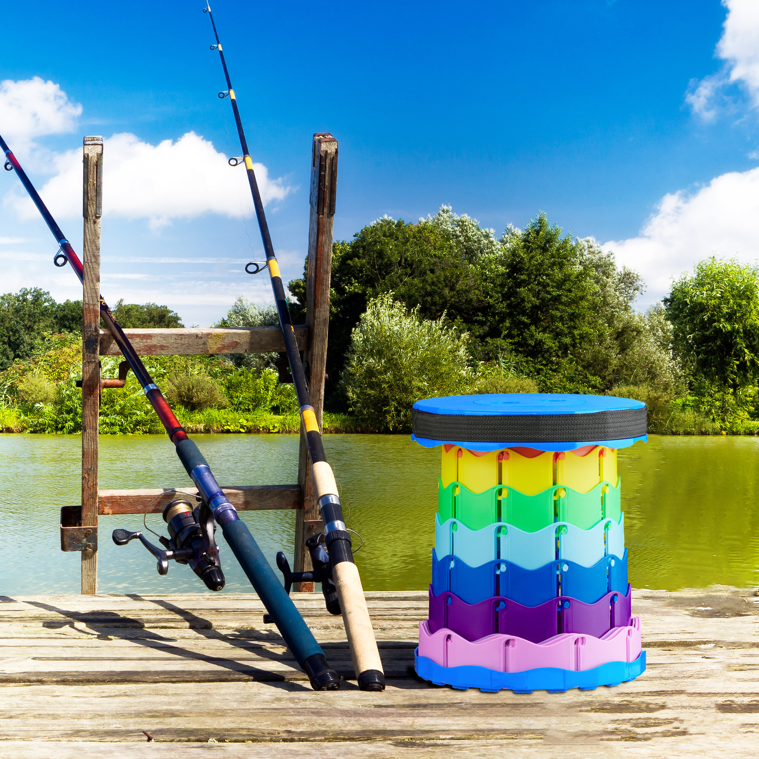 Zwei Angelruten, ein Holzbock und ein bunter Hocker auf einem Holzsteg am See mit Bäumen und blauem Himmel im Hintergrund.