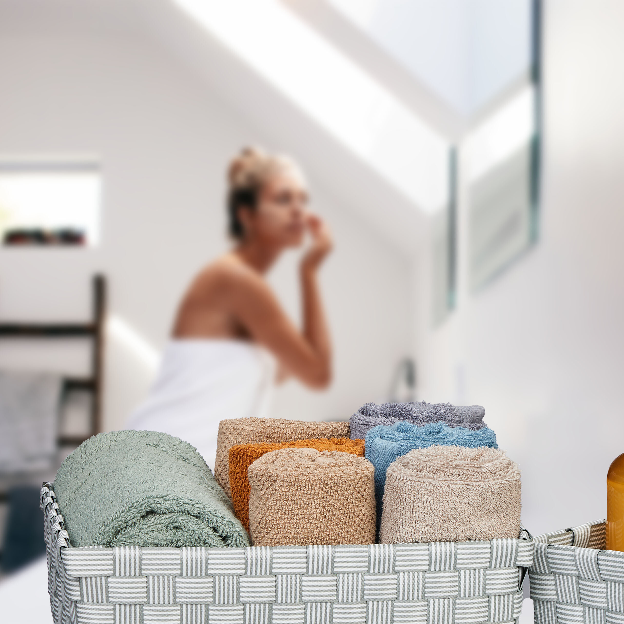 Ringerollte bunte Handtücher in einem grauen Korb, im Hintergrund eine unscharfe Frau im Badezimmer.