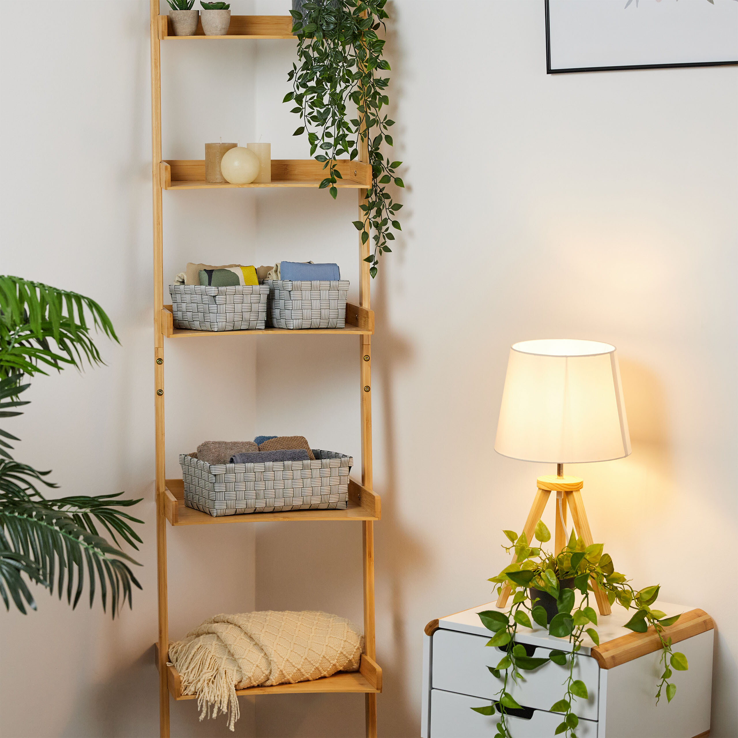 Ein Holzregal mit Korbwaren und Kerzen steht neben einem Nachttisch mit einer Lampe und einer Pflanze.