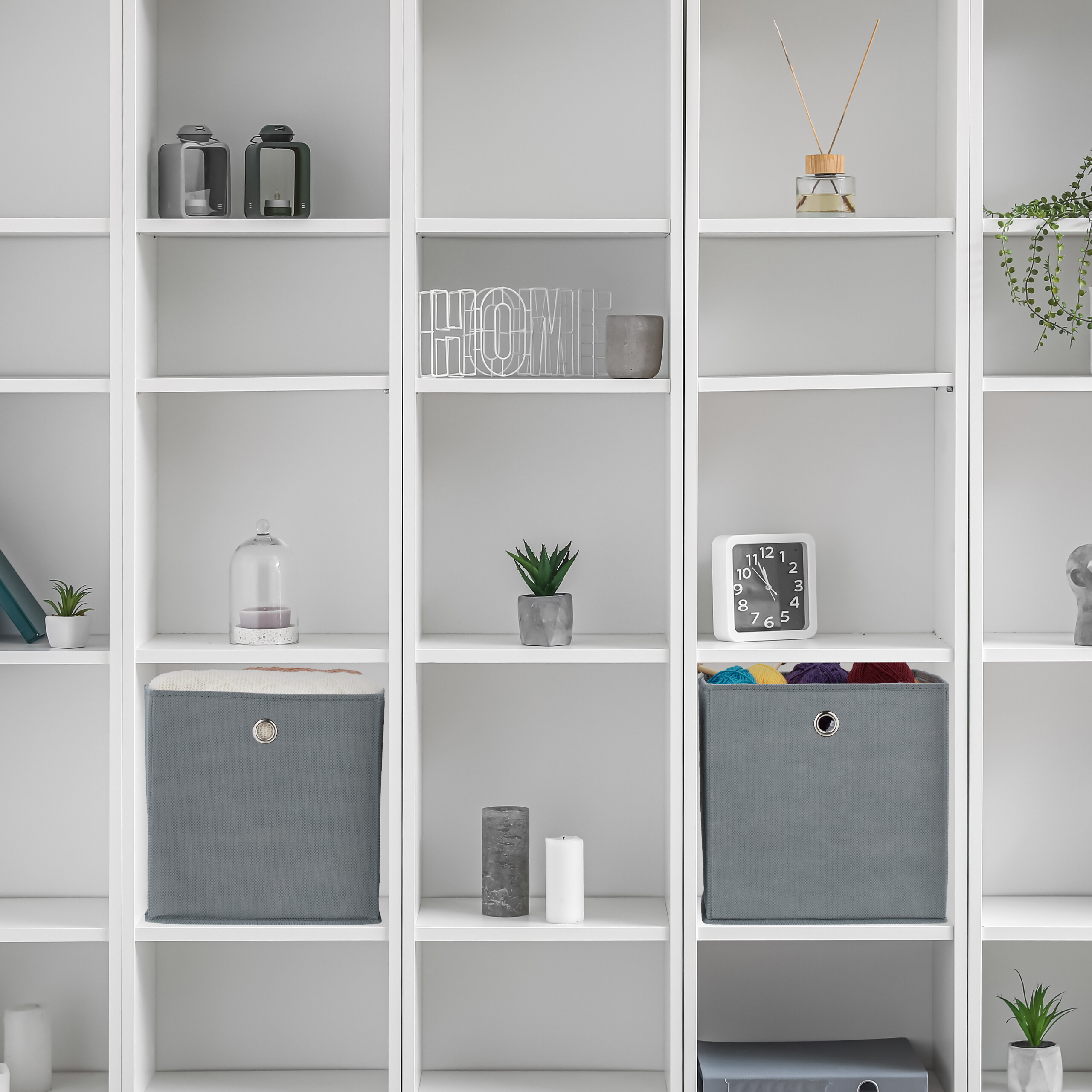 Ein weißes modulares Regal mit mehreren Fächern enthält Dekoartikel, Pflanzen, eine Uhr und Aufbewahrungsboxen.