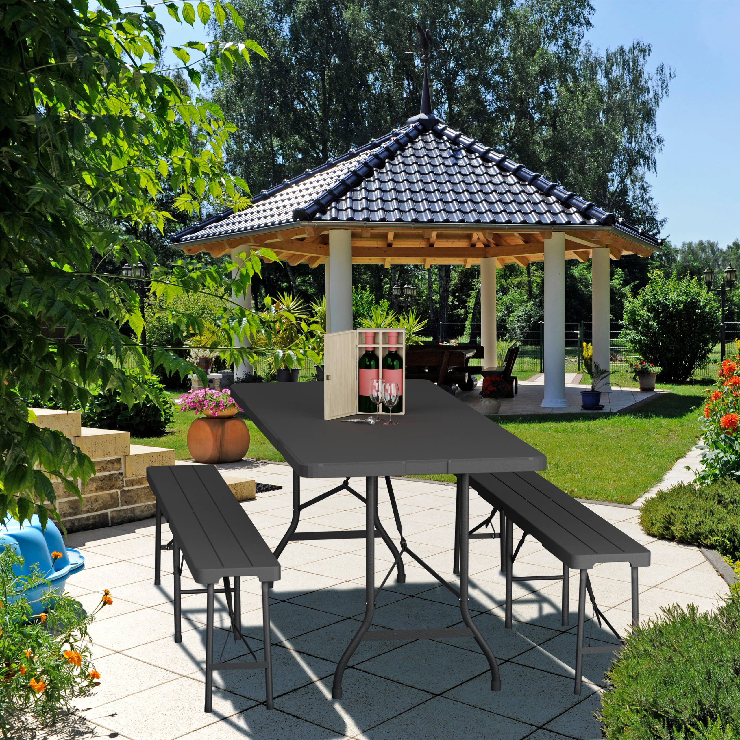 Ein grauer Gartentisch mit zwei Bänken steht auf einer Terrasse vor einem Pavillon und grünem Garten.