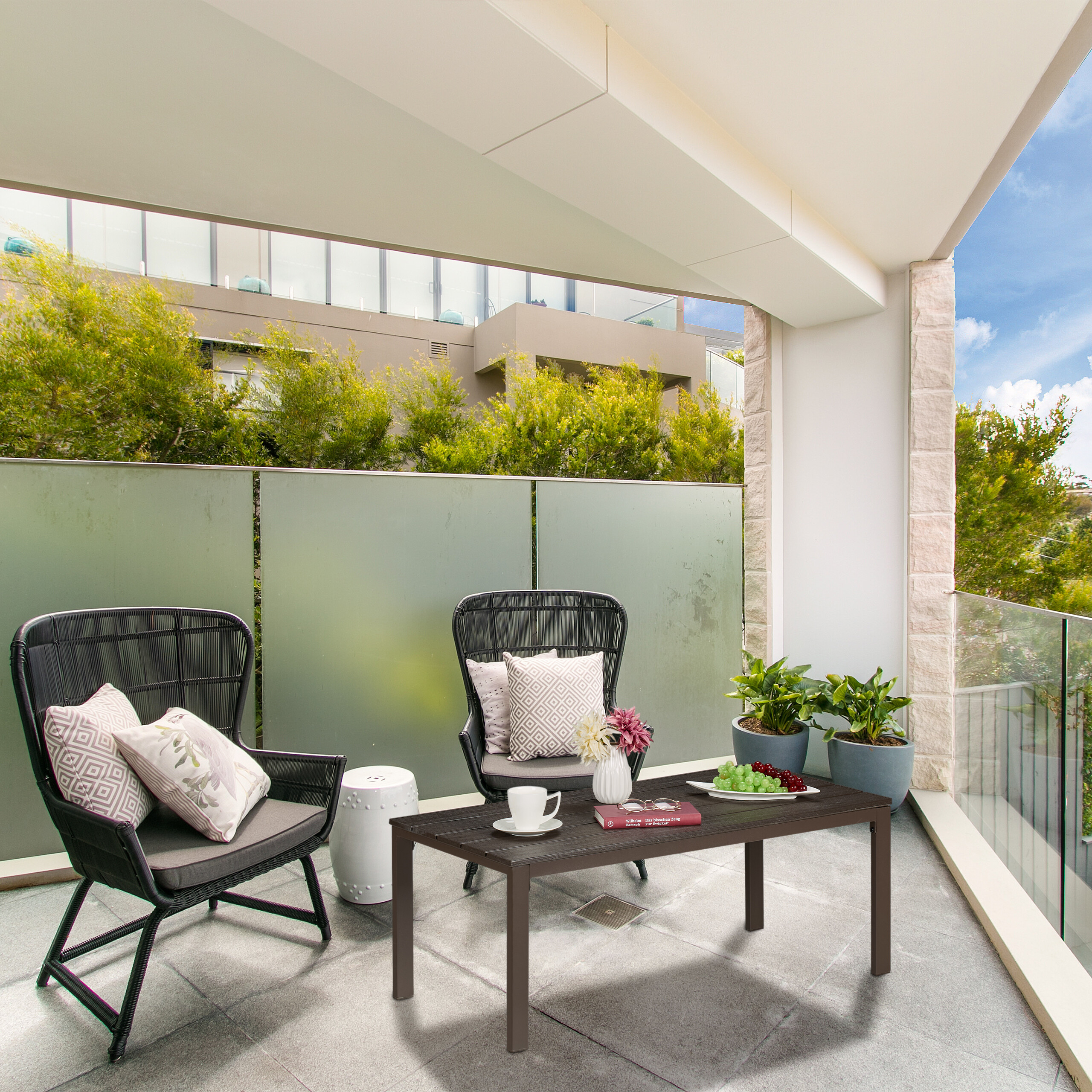 Moderne Balkonmöbel mit Sesseln, Tisch und Kissen vor einer Glasbrüstung mit Bäumen im Hintergrund.