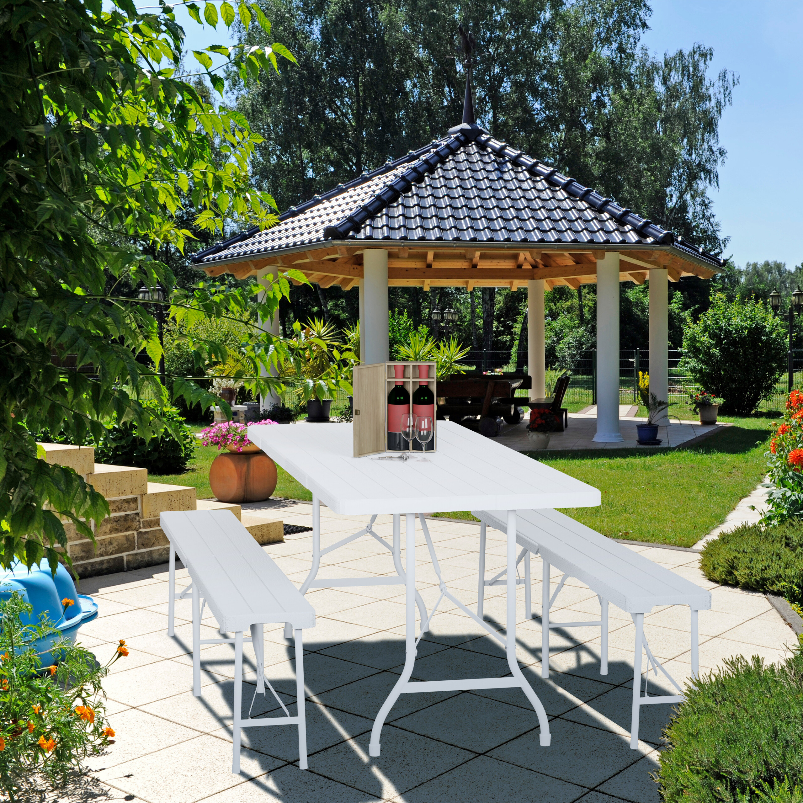 Ein weißer Klapptisch mit zwei Bänken steht auf einer Terrasse im Garten, im Hintergrund ein offener Pavillon.