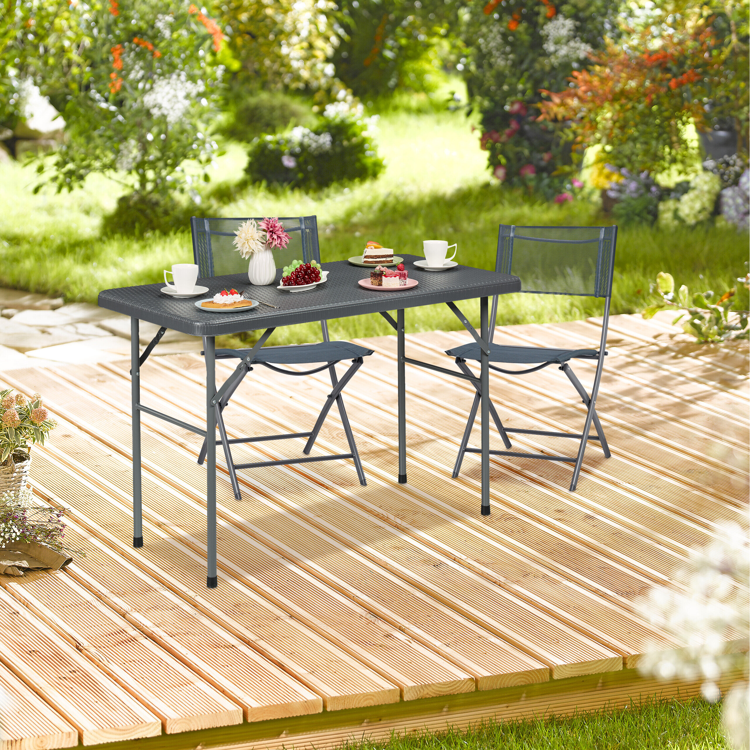 Ein grauer Gartentisch mit zwei Stühlen auf einer Holzterrasse im Garten, gedeckt mit Kuchen, Obst und Tassen.