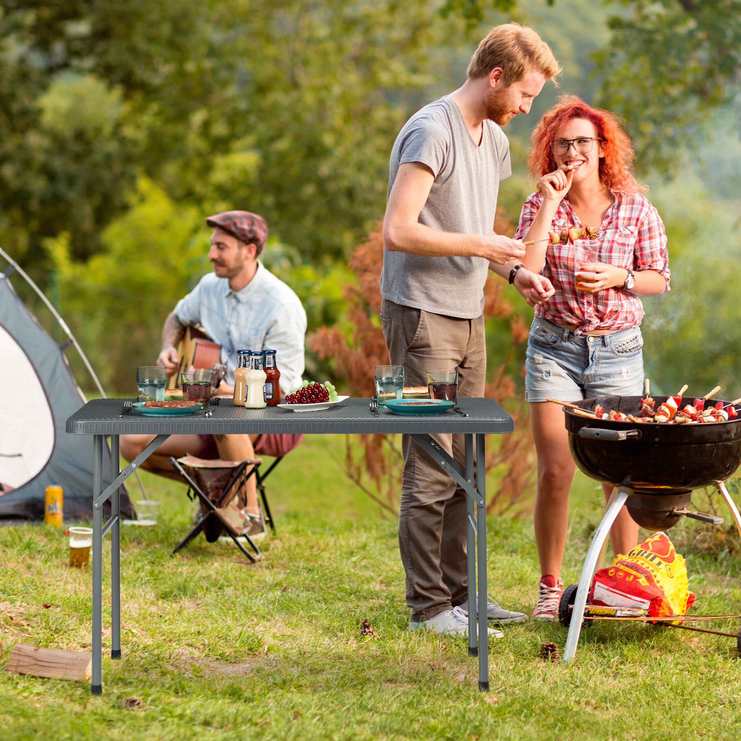Freunde beim Camping: Ein Paar grillt im Freien, während ein anderer Mann Gitarre spielt und auf dem Tisch Essen steht.