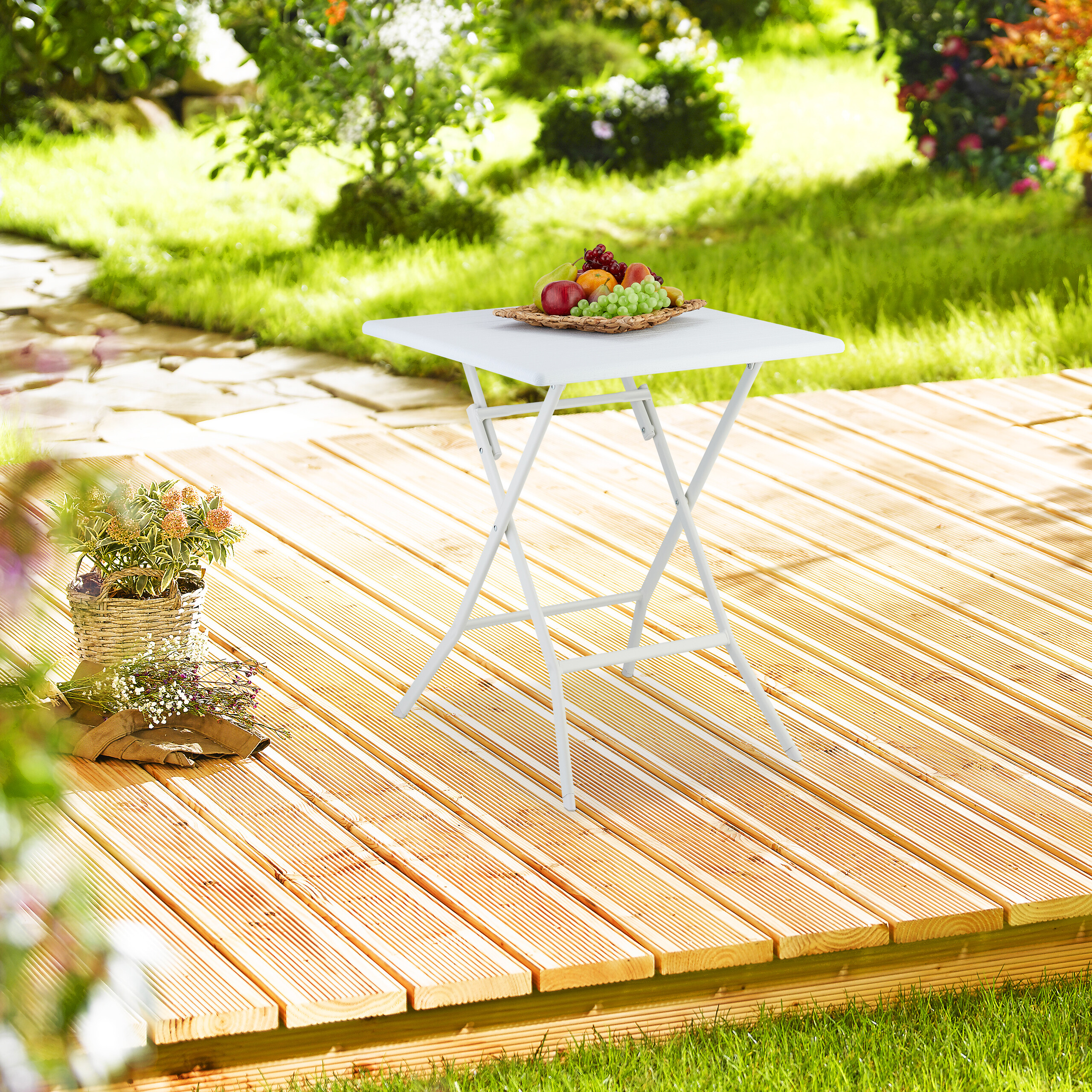 Ein weißer Klapptisch mit Früchten steht auf einer Holzterrasse im sonnigen Garten.