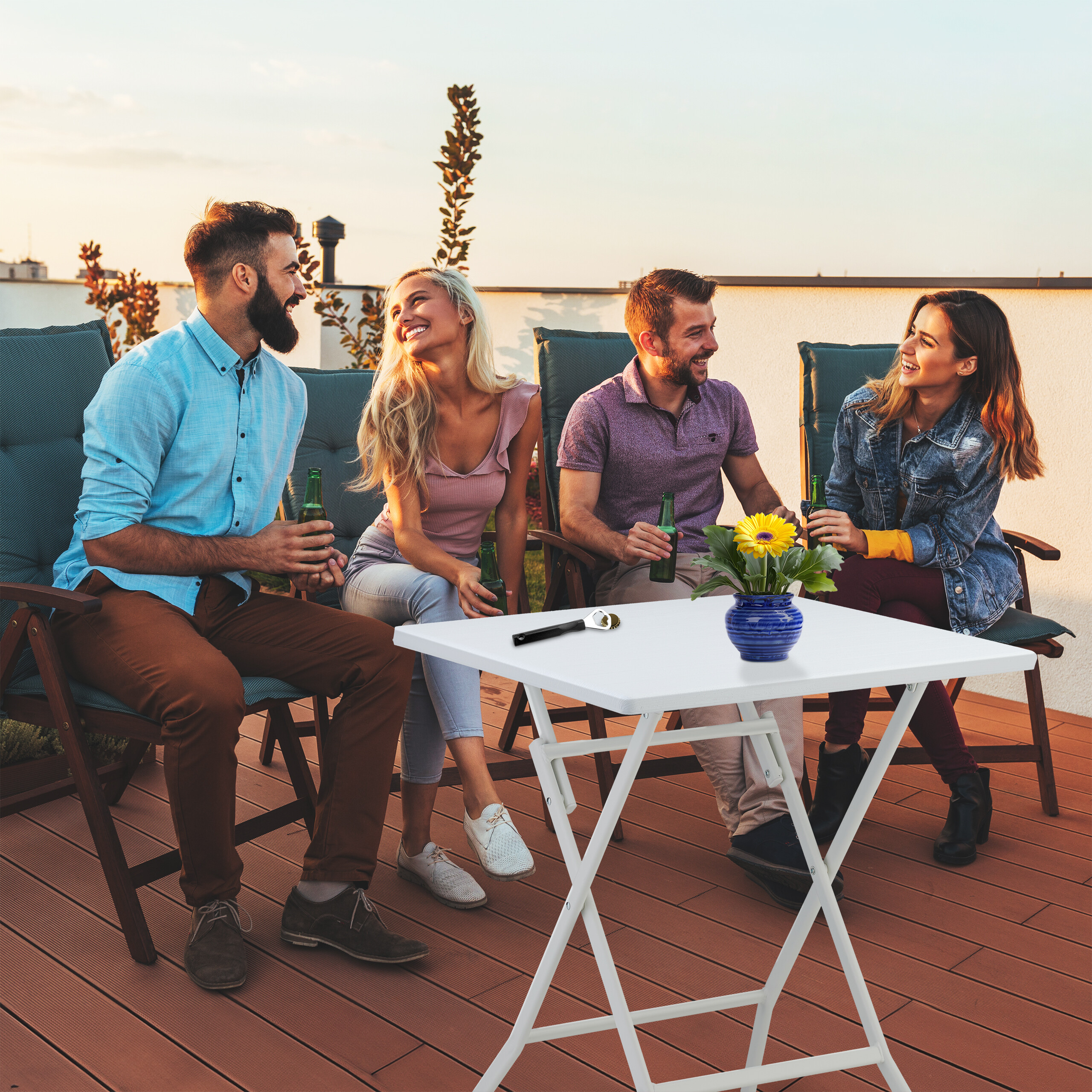 Vier Freunde sitzen bei Sonnenuntergang auf einer Dachterrasse, halten Getränke und unterhalten sich an einem Tisch.