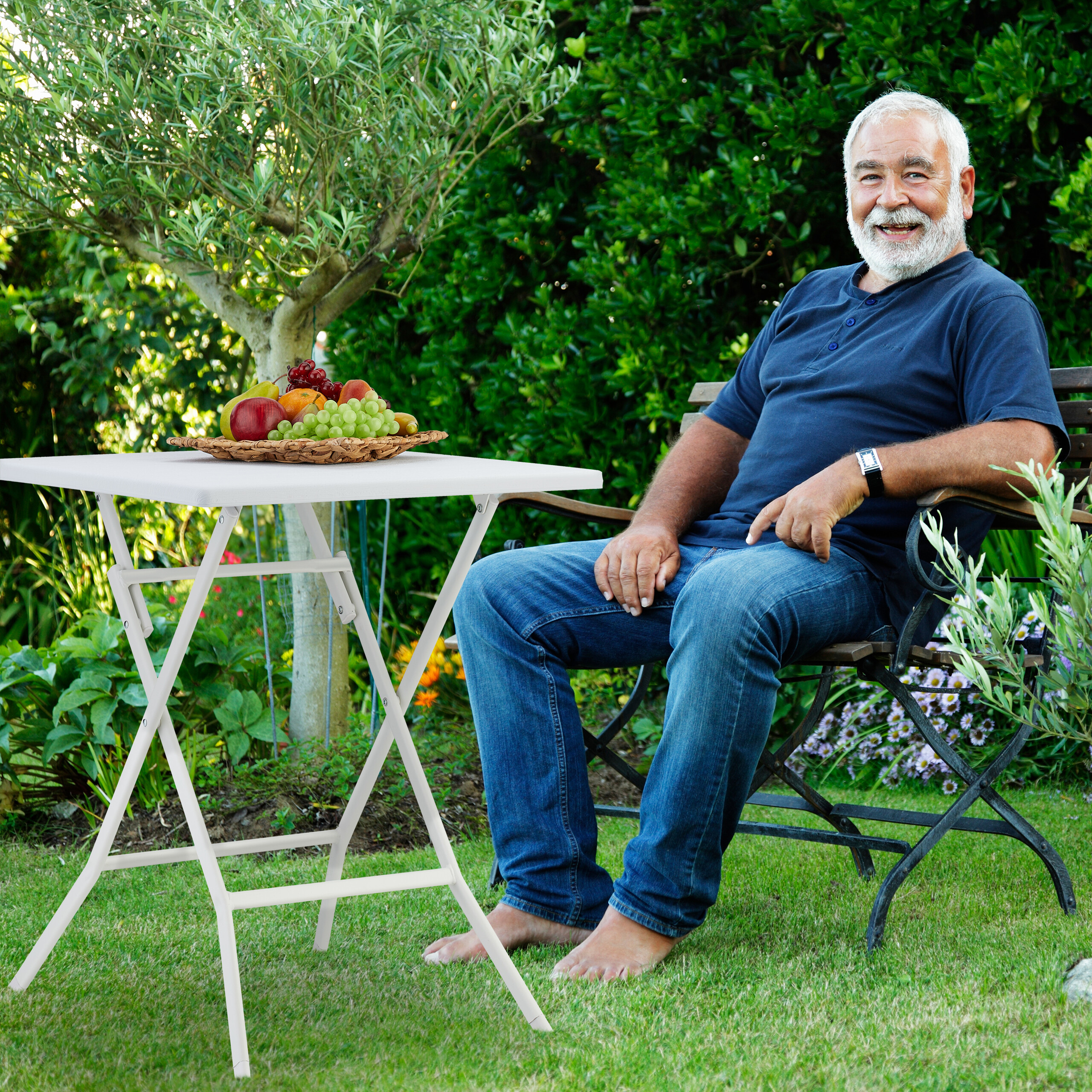 Ein älterer Mann mit grauem Bart sitzt im Garten auf einer Bank neben einem Tisch mit Früchten.
