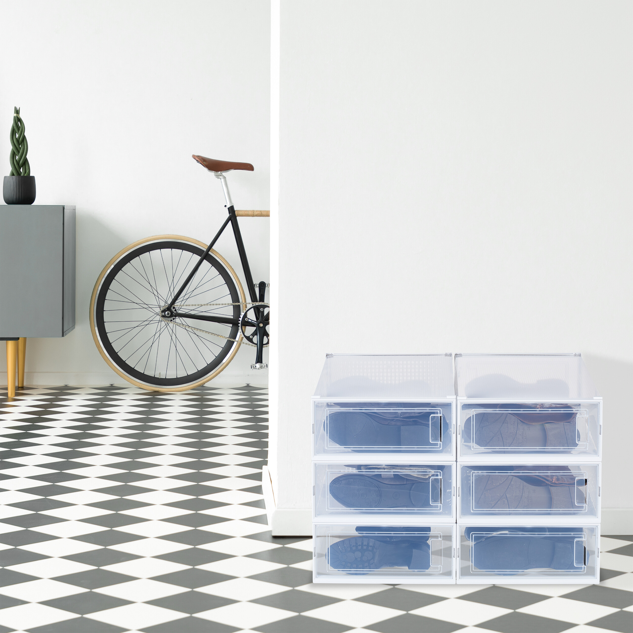Sechs transparente Schuhkartons stehen auf einem schwarz-weiß gefliesten Boden neben einer weißen Wand und einem Fahrrad.
