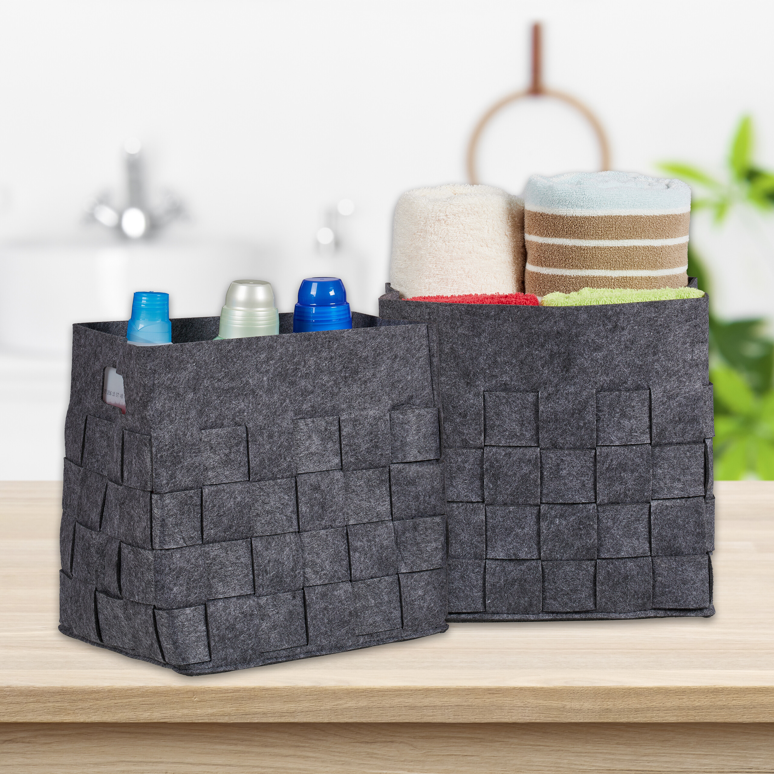 Zwei graue Filzkörbe mit Flaschen und Handtüchern auf einem Holztisch vor einem verschwommenen Badezimmer.