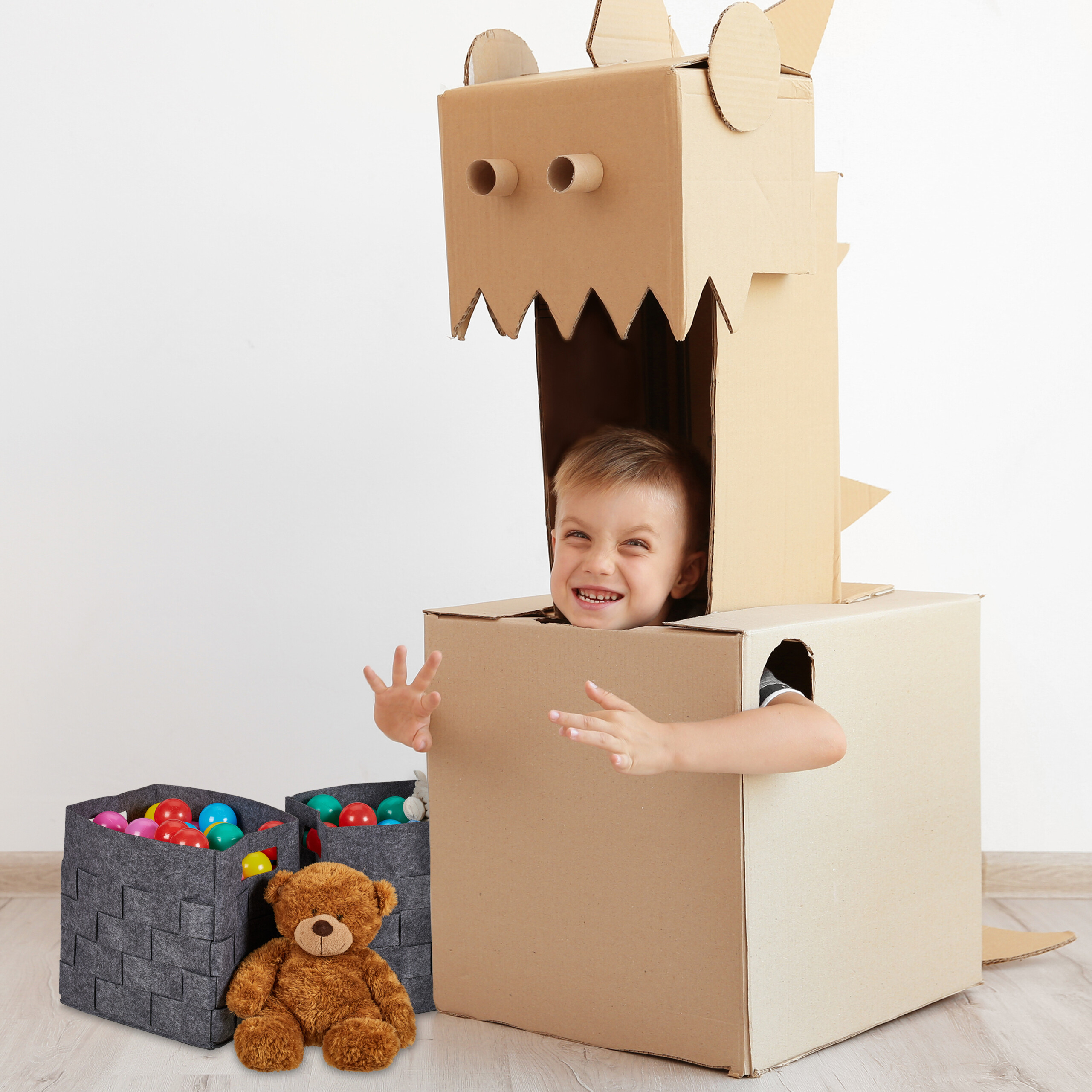 Ein glücklicher Junge in einem selbstgemachten Pappdrachenkostüm lächelt, neben ihm ein Teddybär und bunte Bälle.