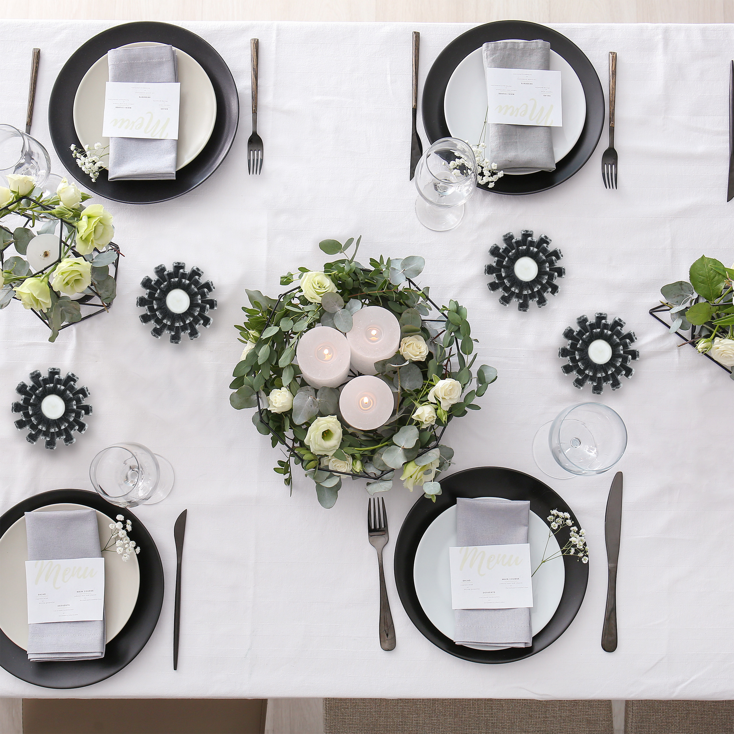 Draufsicht auf einen eleganten Esstisch mit Gedecken, Blumengestecken und brennenden Kerzen auf weißem Tuch.