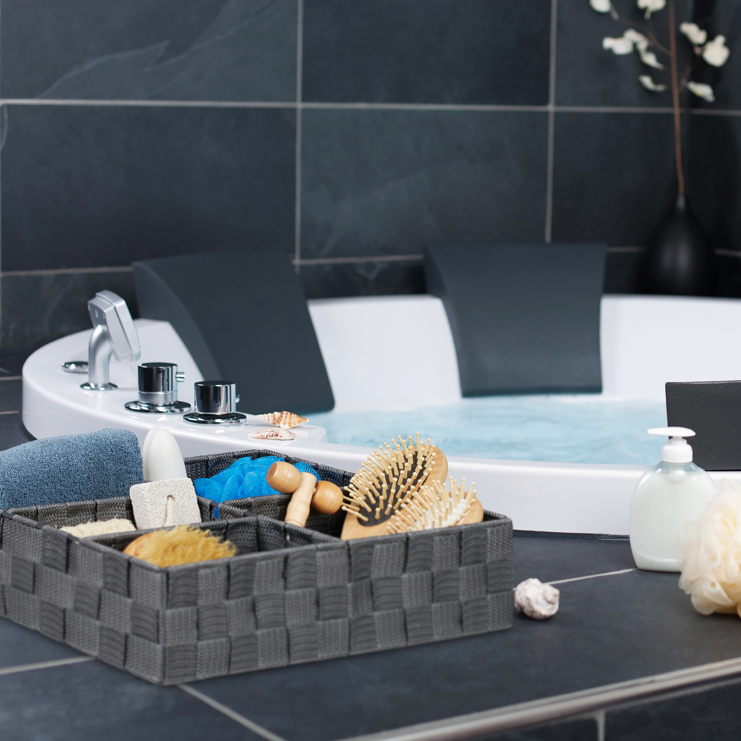 Ein grauer Aufbewahrungskorb mit Badartikeln steht auf dunklen Fliesen vor einer weißen Whirlpool-Badewanne.