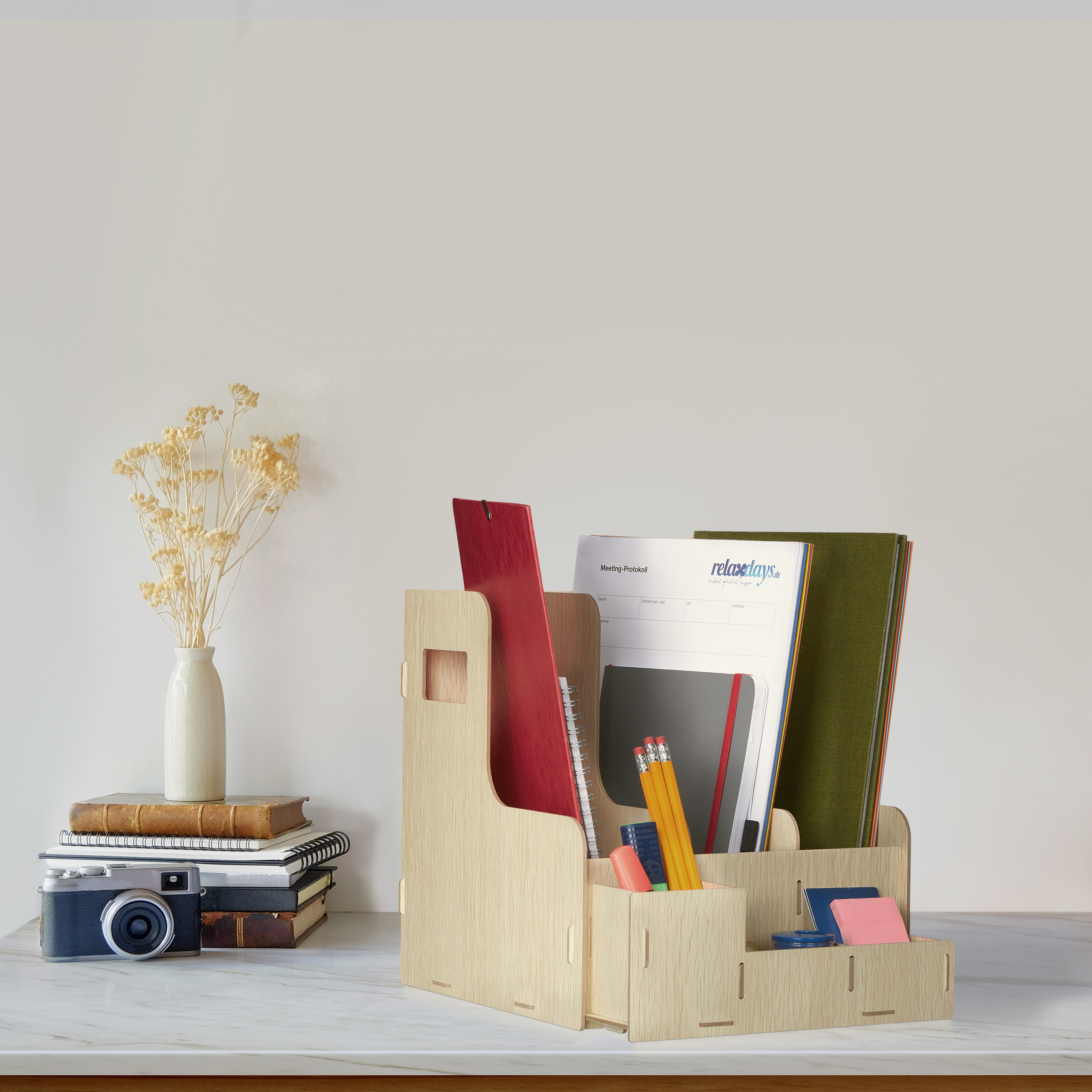 Ein hölzerner Dokumentenorganizer mit Notizbüchern und Stiften steht auf einem Tisch neben einer Kamera und Blumen.
