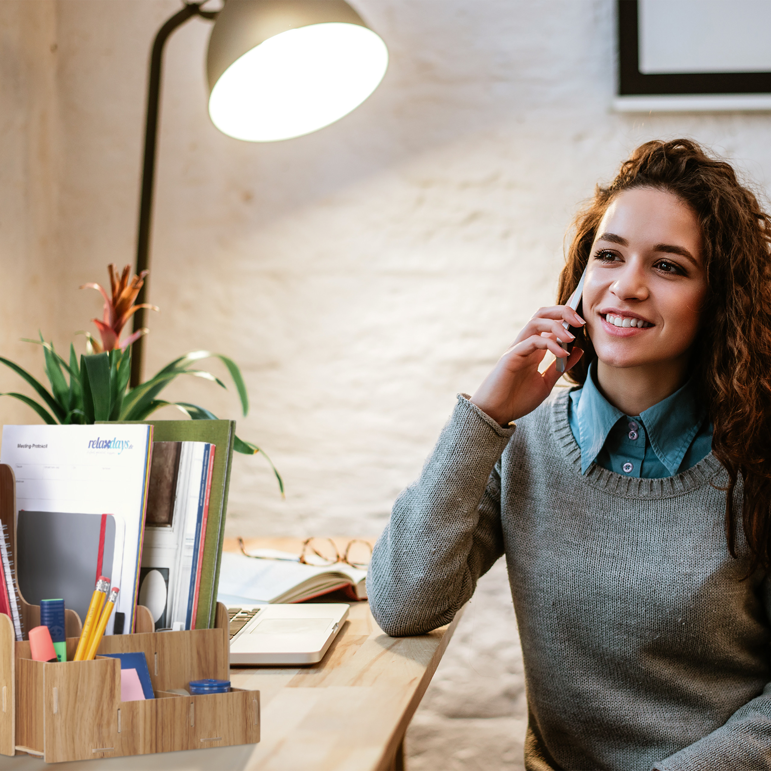 Eine junge Frau mit lockigem Haar telefoniert am Holztisch mit Lampe und Dokumenten.