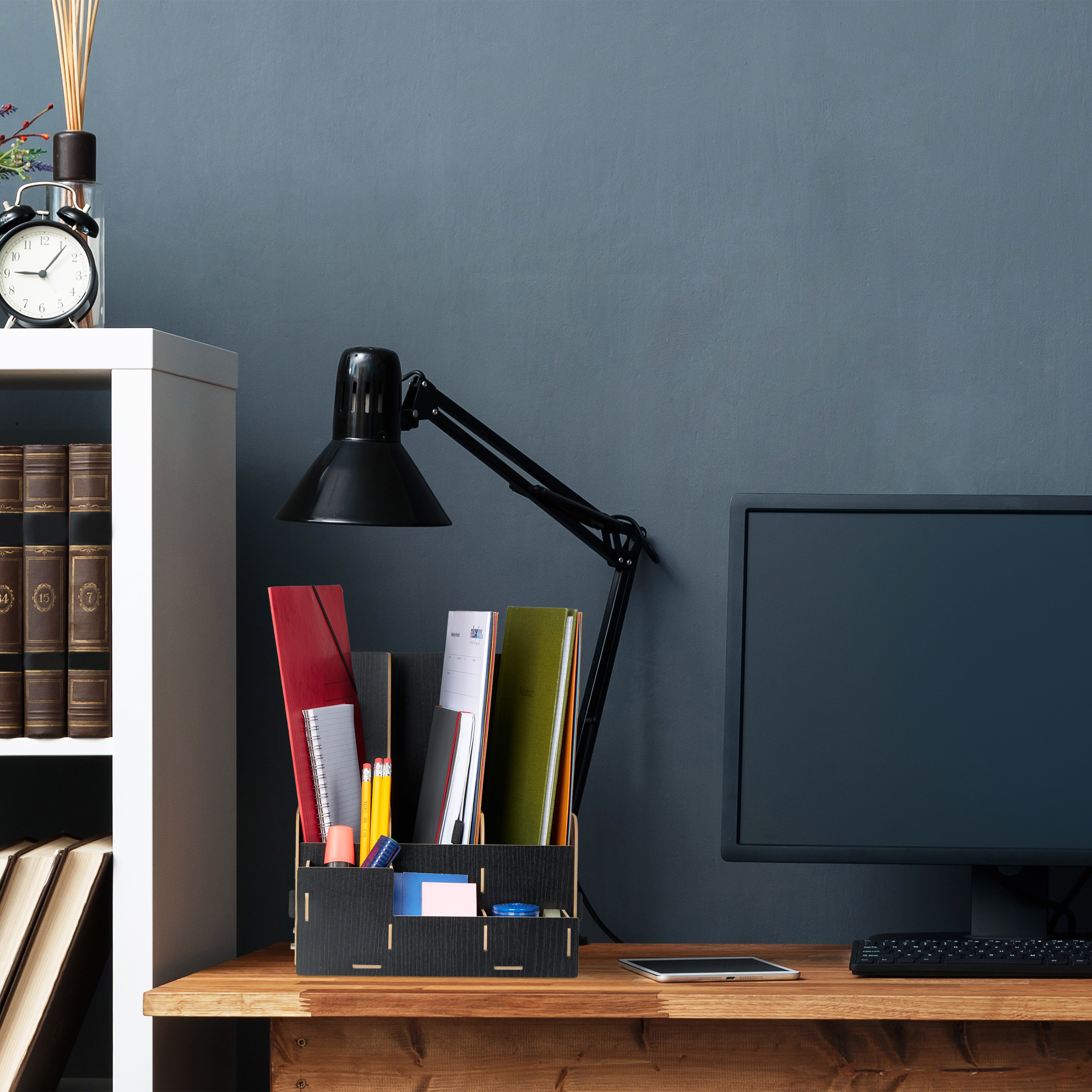 Ein Schreibtisch mit Monitor, Lampe, Organizer voller Ordner, Stifte und ein Regal vor grauer Wand.