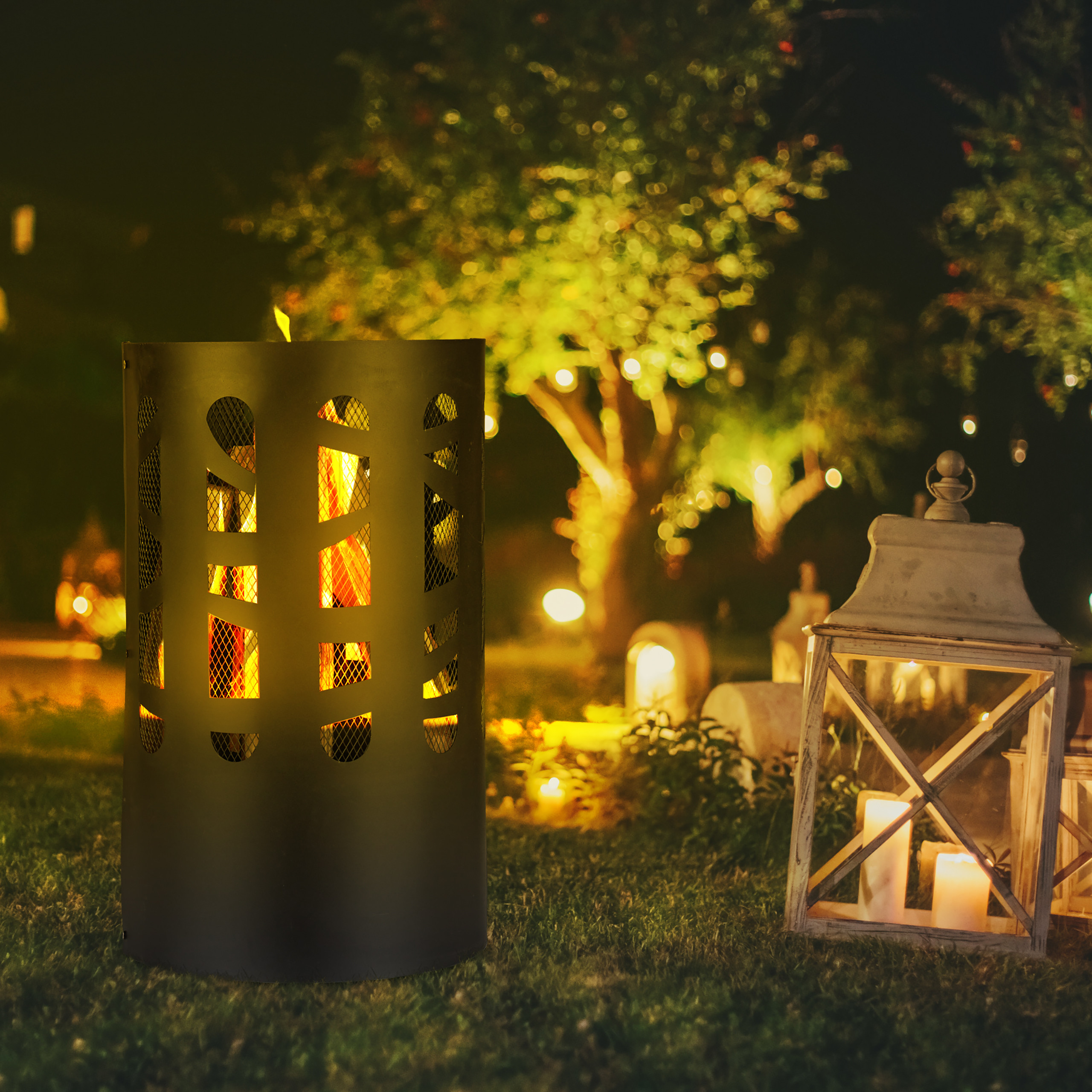 Eine schwarze Feuersäule und eine weiße Kerzenlaterne stehen nachts auf dem Rasen im beleuchteten Garten.