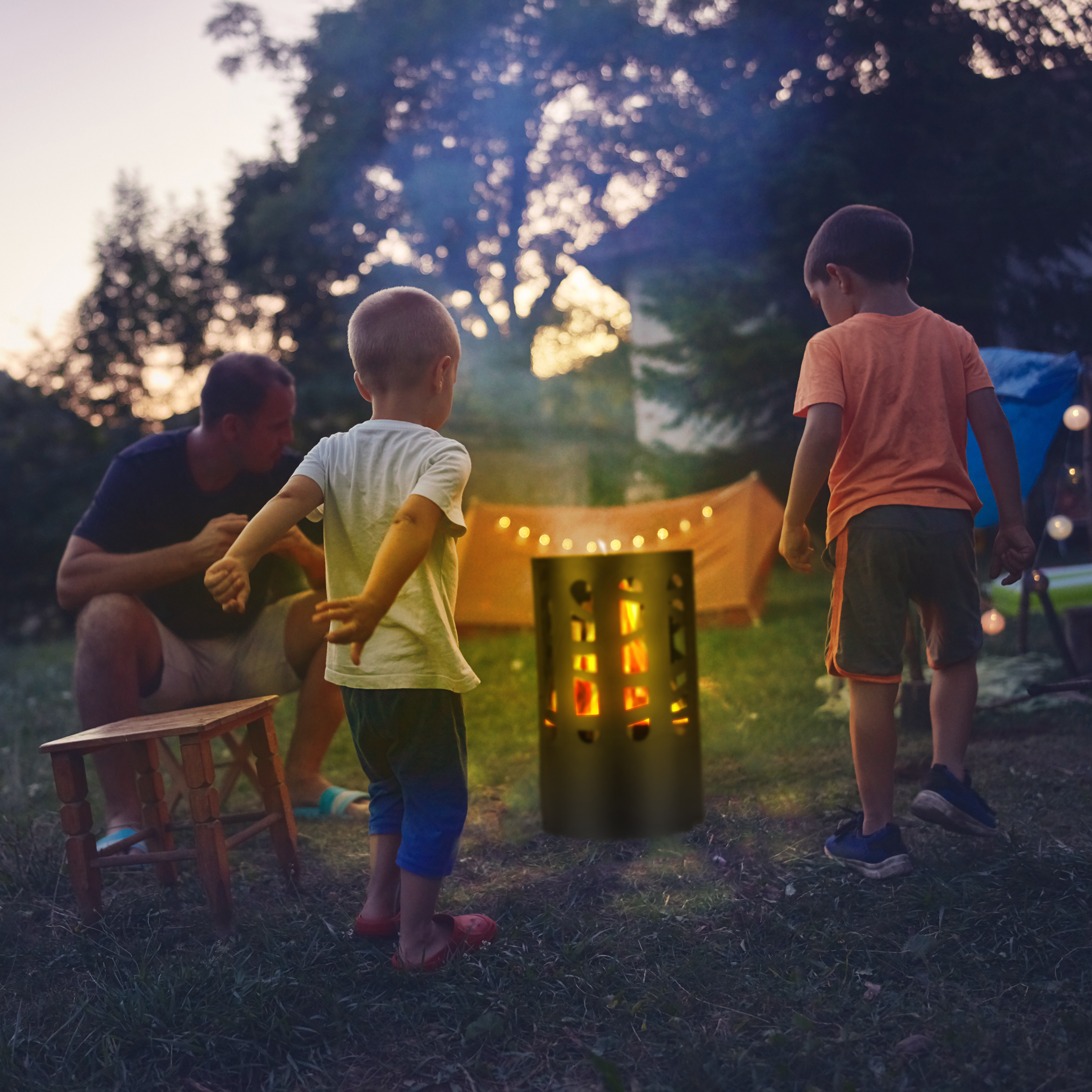 Ein Mann und zwei Jungen campen im Garten bei einem kleinen Feuer in einem zylindrischen Behälter.