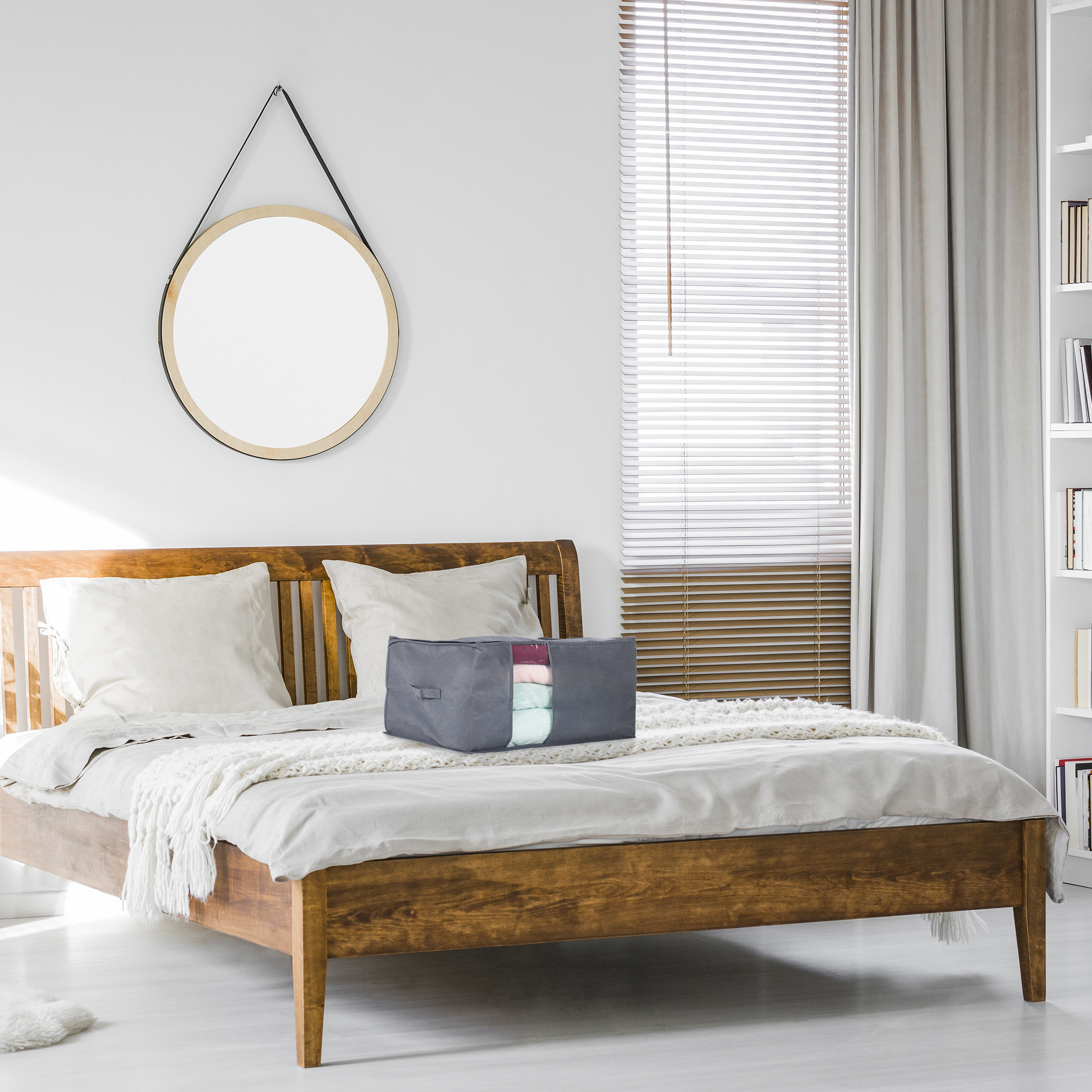 Eine graue Stoffaufbewahrungstasche mit gefalteter Kleidung liegt auf einem Holzbett in einem hellen Schlafzimmer.
