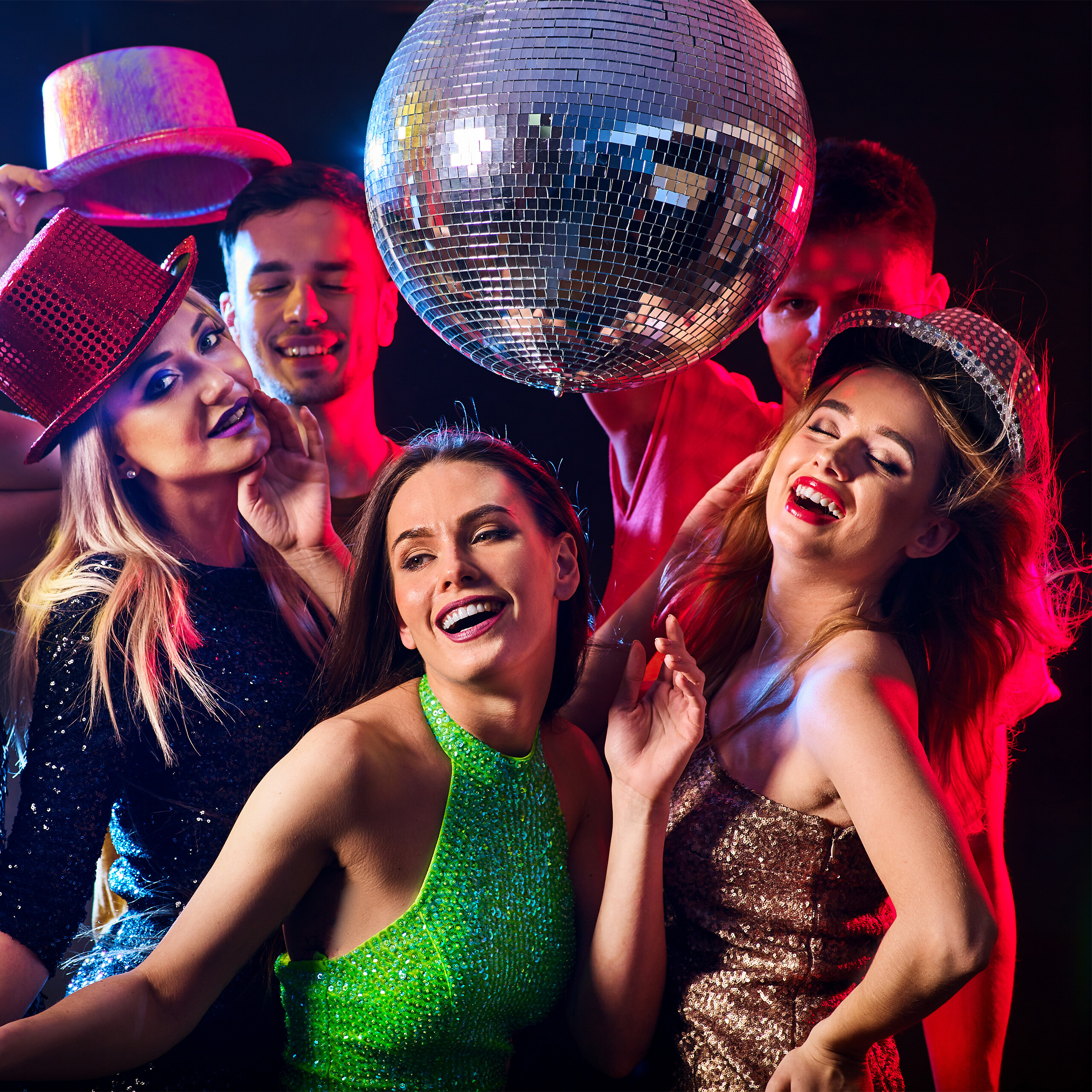 Freunde in Partyhüten tanzen fröhlich unter einer Discokugel bei bunter Beleuchtung.