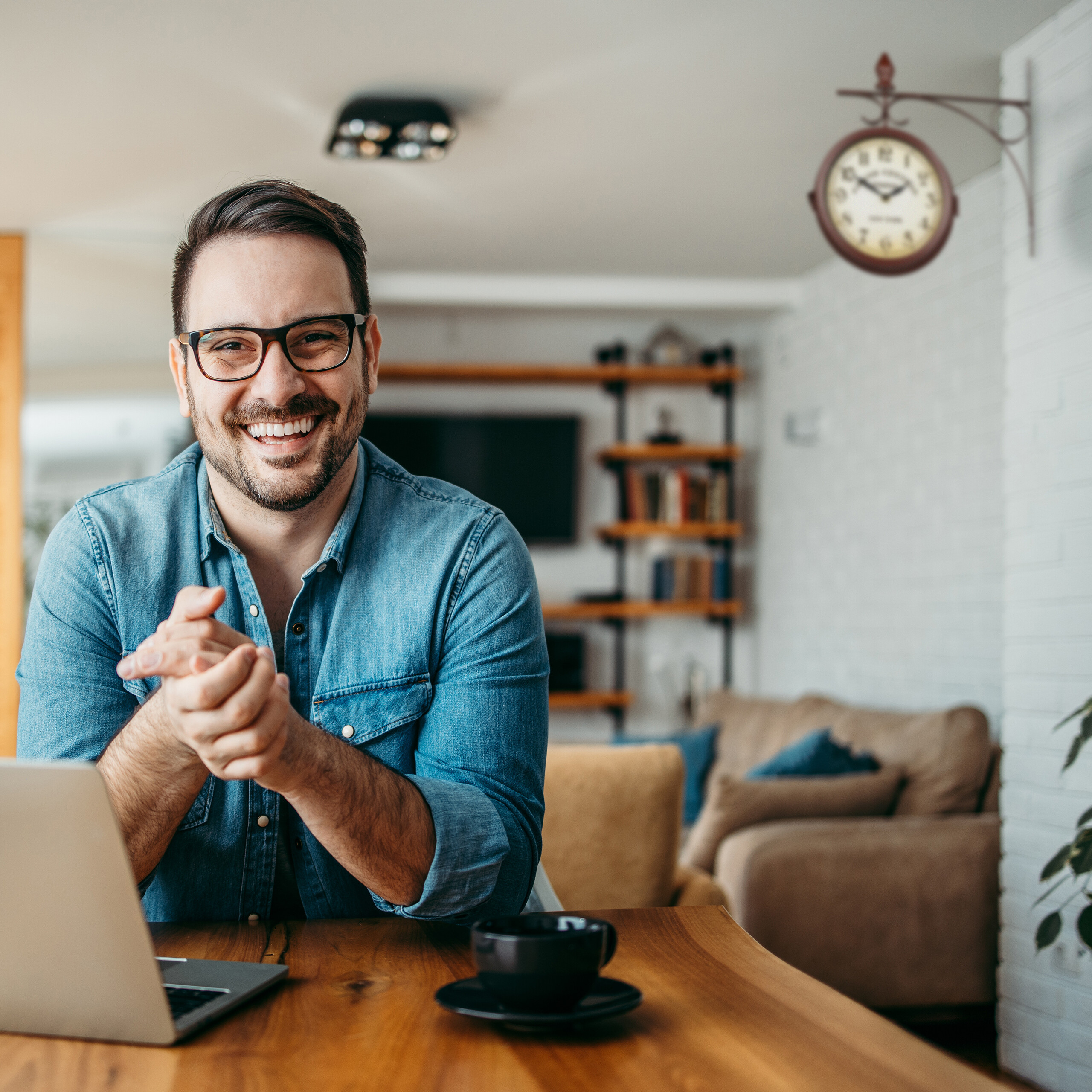 Lächelnder Mann mit Brille und Jeanshemd sitzt an Holztisch mit Laptop und Kaffeetasse.