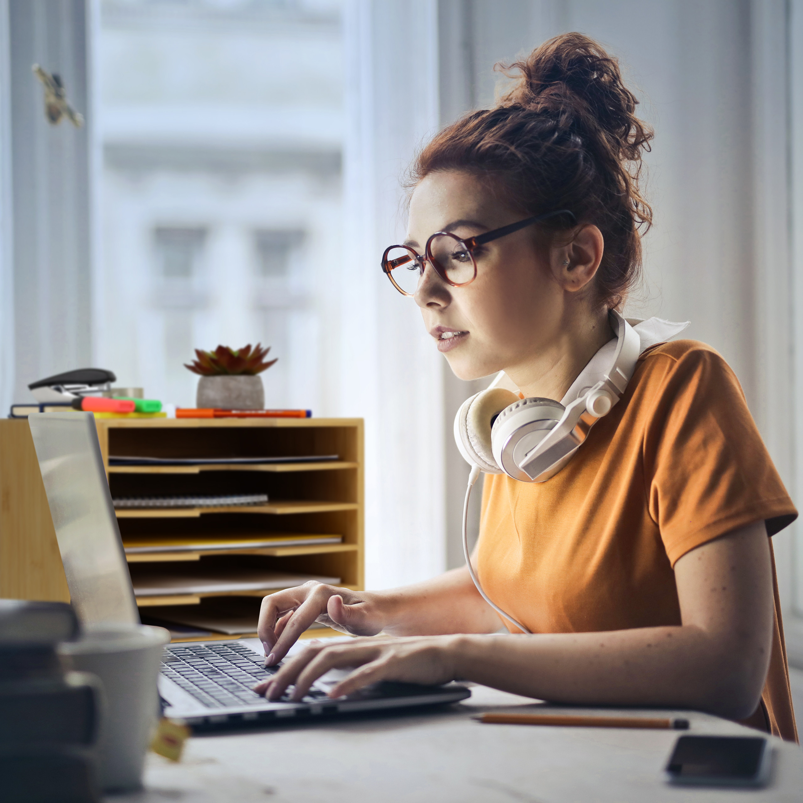 Eine Frau mit Brille und Kopfhörern tippt in einem hellen Raum auf einen Laptop.