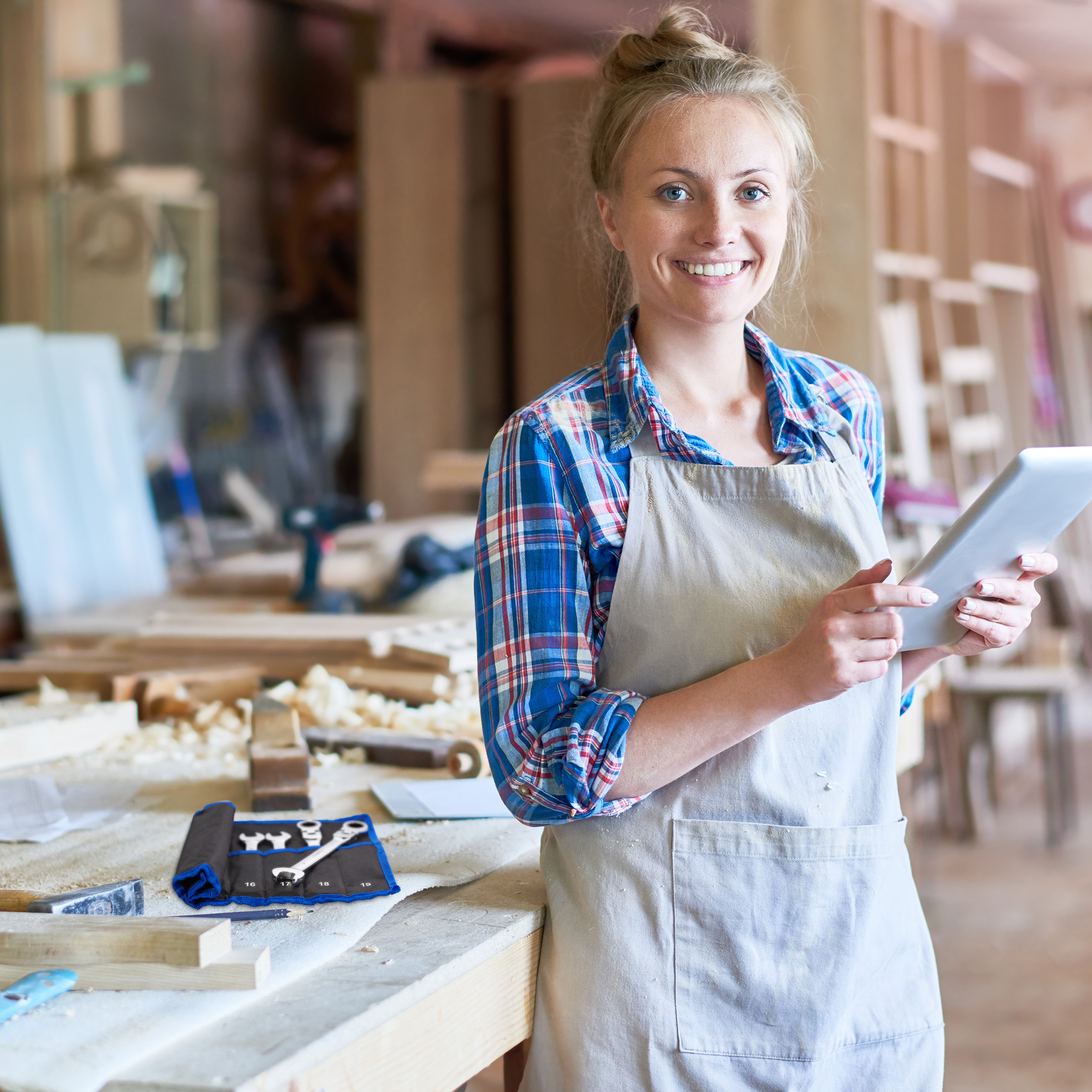 Eine Handwerkerin mit Schürze hält ein Tablet in einer Werkstatt.