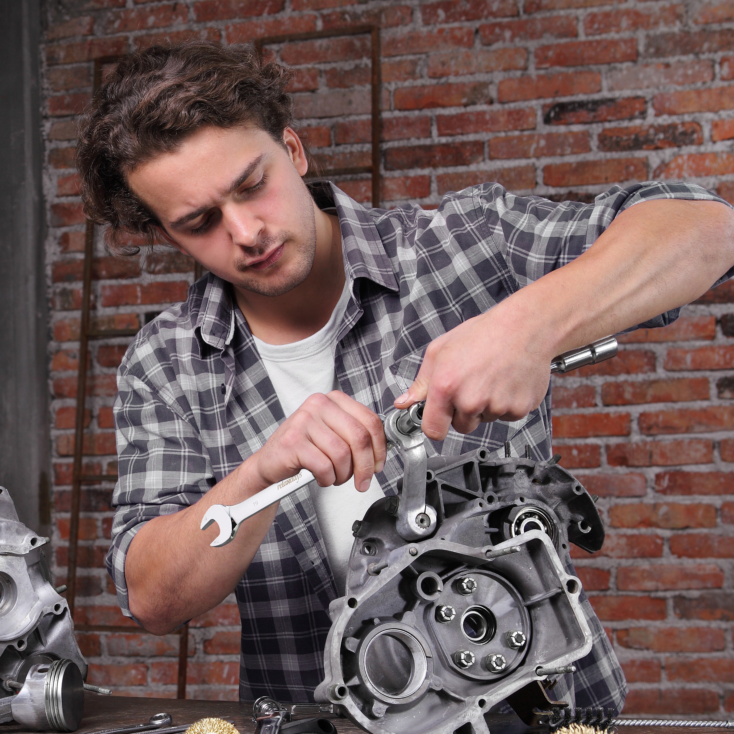 Ein Mann im karierten Hemd repariert einen Motor mit Werkzeugen vor einer Ziegelwand.