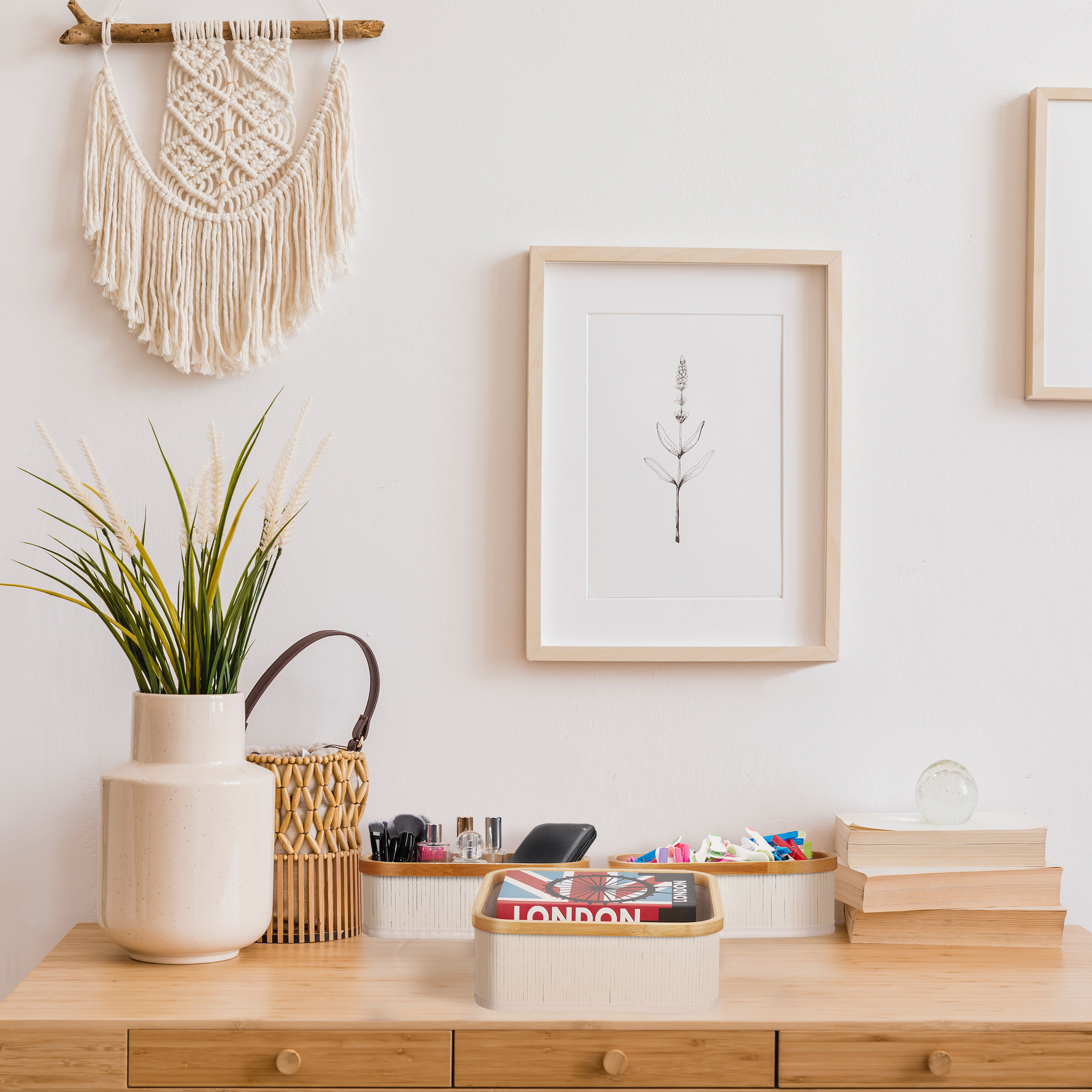 Ein Holztisch zeigt eine weiße Vase, gerahmte Pflanzenkunst, Körbe, Bücher und einen Makramee-Wandbehang.