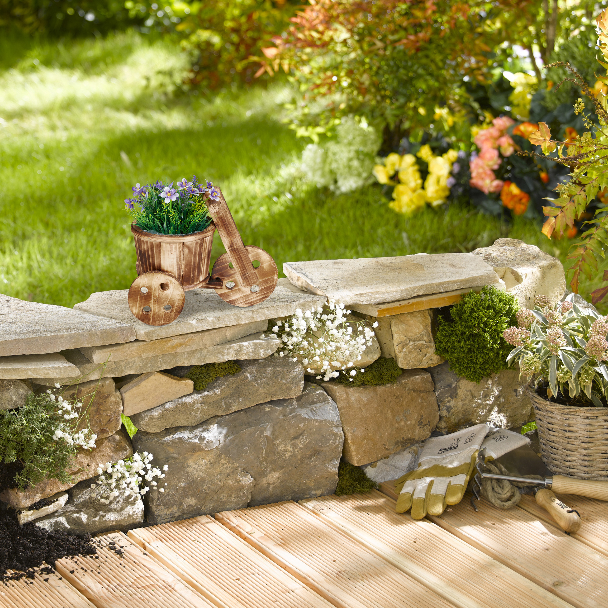 Holzkübel mit lila Blumen auf einer Steinmauer im garten, dazu Gartenhandschuhe und Werkzeug auf Holzboden.