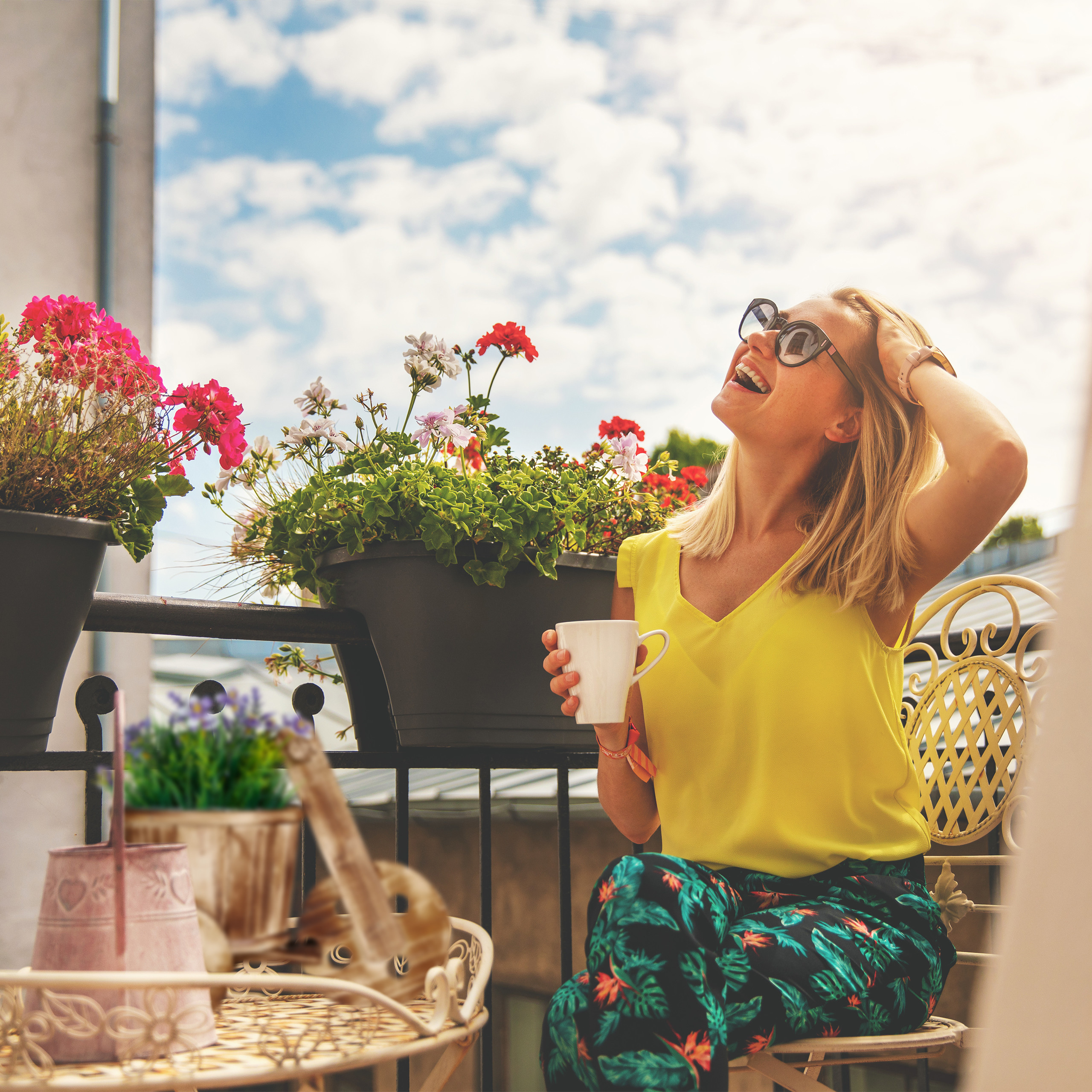 Eine blonde Frau auf einem sonnigen Balkon mit Kaffeetasse und Blumen.