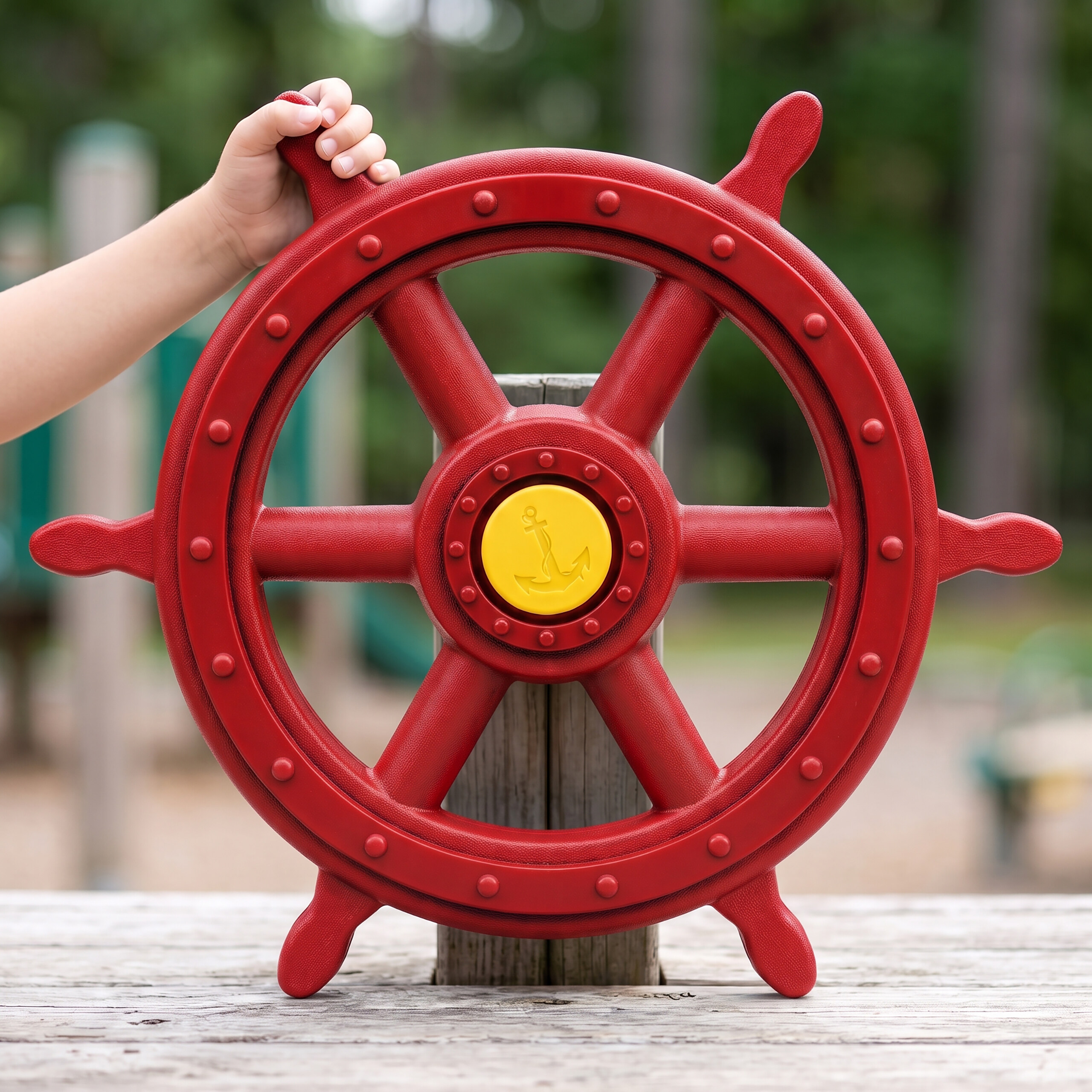 Die Hand eines Kindes hält ein großes rotes Schiffssteuerrad mit einem gelben Anker auf einem Spielplatz.