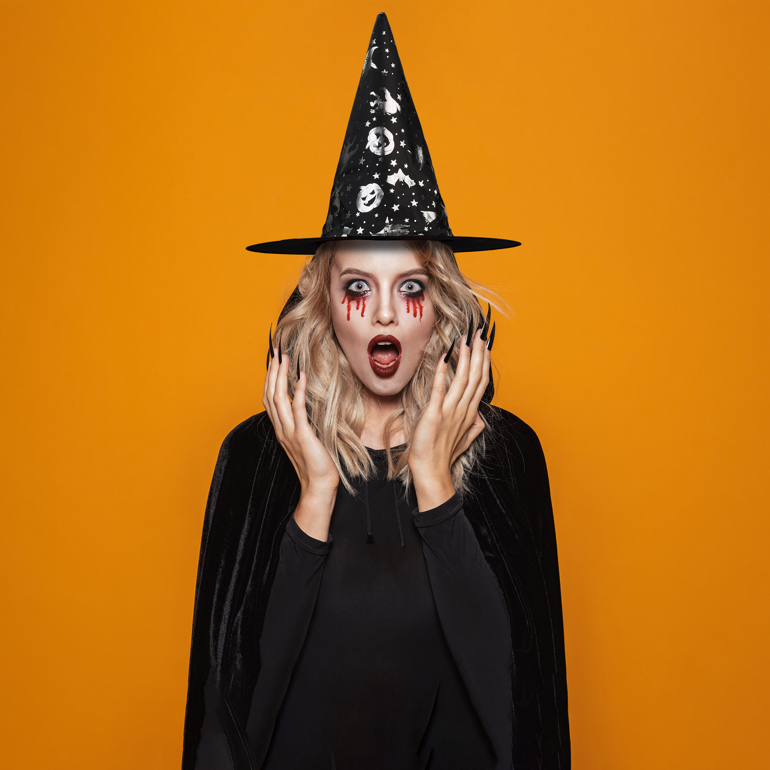 Eine Frau in Hexenkostüm mit schwarzem Hut, rotem Make-up und Cape vor gelbem Hintergrund.