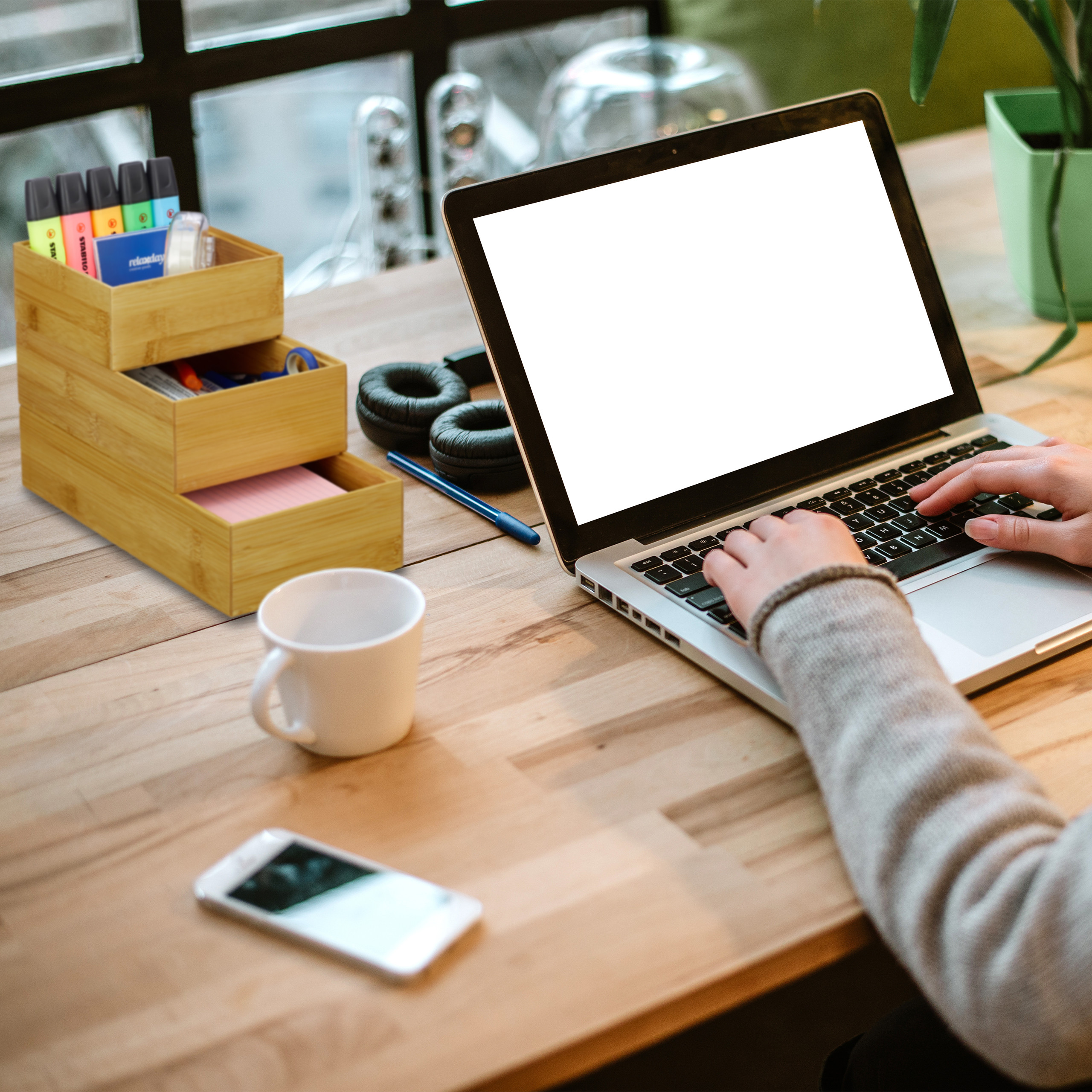 Person tippt auf Laptop am Holztisch mit Becher, Stiften und Smartphone.
