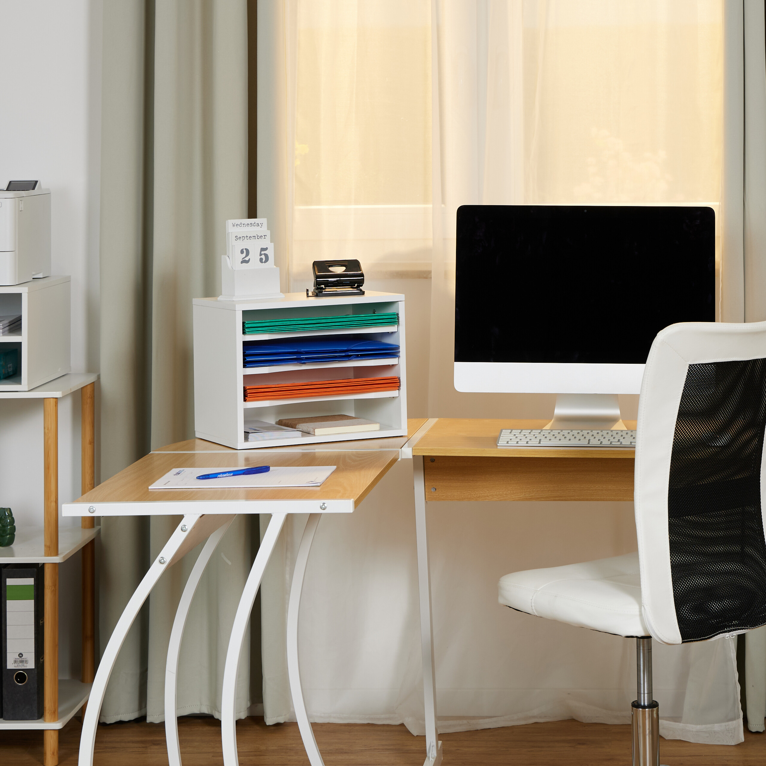 Ein heller Schreibtisch mit Computer, Dokumentenorganizer, Kalender und weißem Stuhl vor einem Fenster mit Vorhängen.