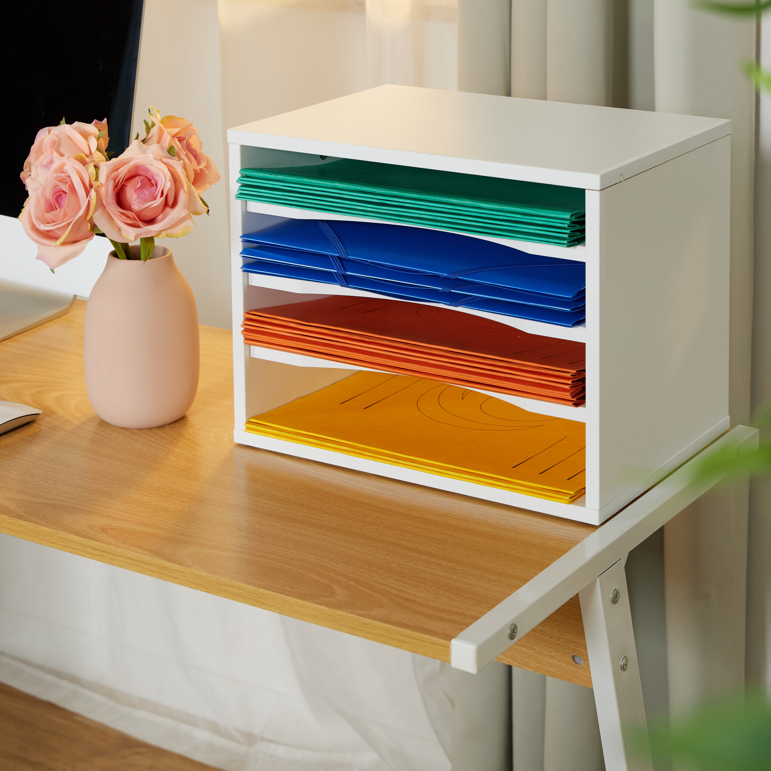Ein weißer Dokumentenorganizer auf einem Holztisch mit bunten Mappen und einer rosa Vase mit Rosen.