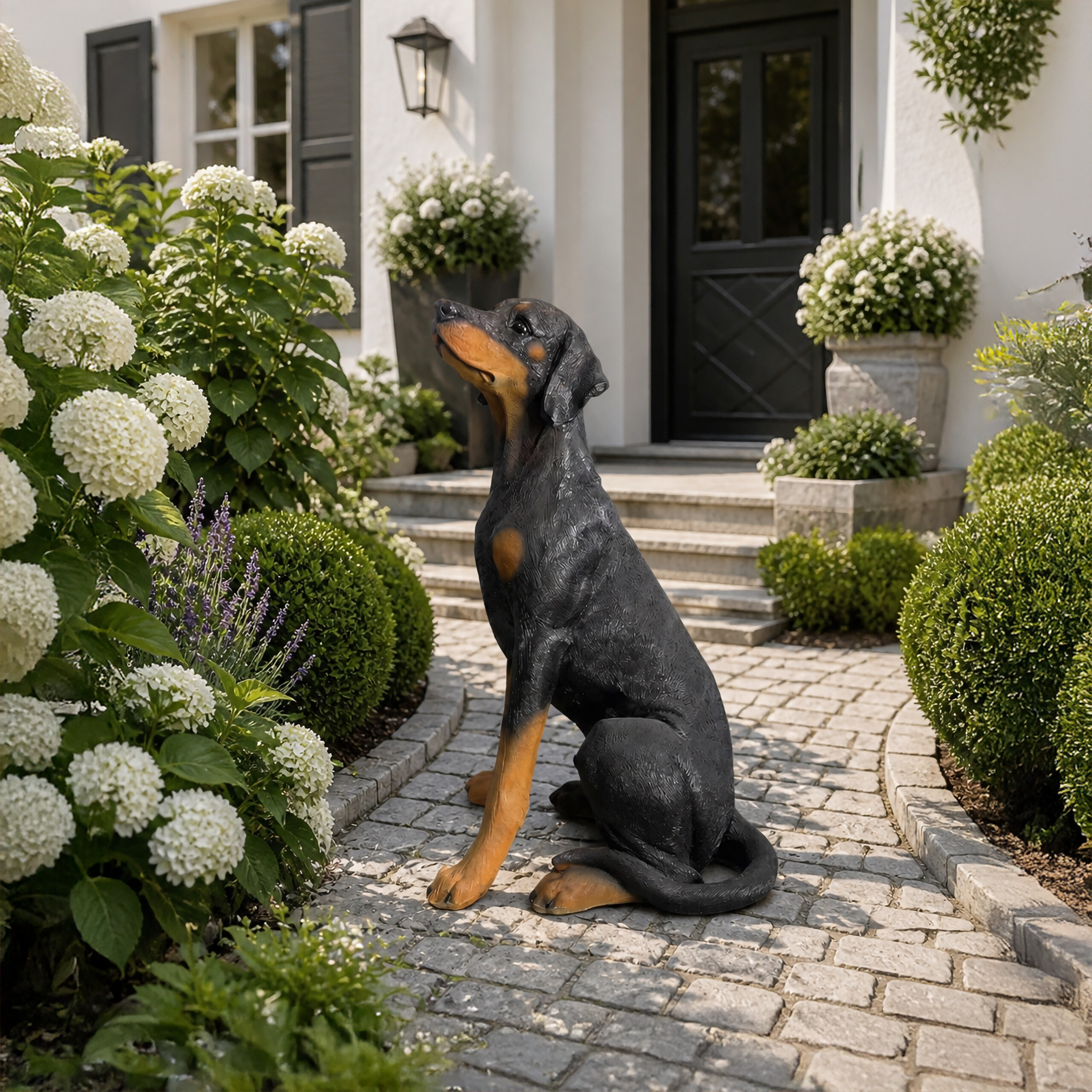 Eine Dobermann-Statue sitzt auf einem Steinweg vor einem Haus, umgeben von weißen Blumen.