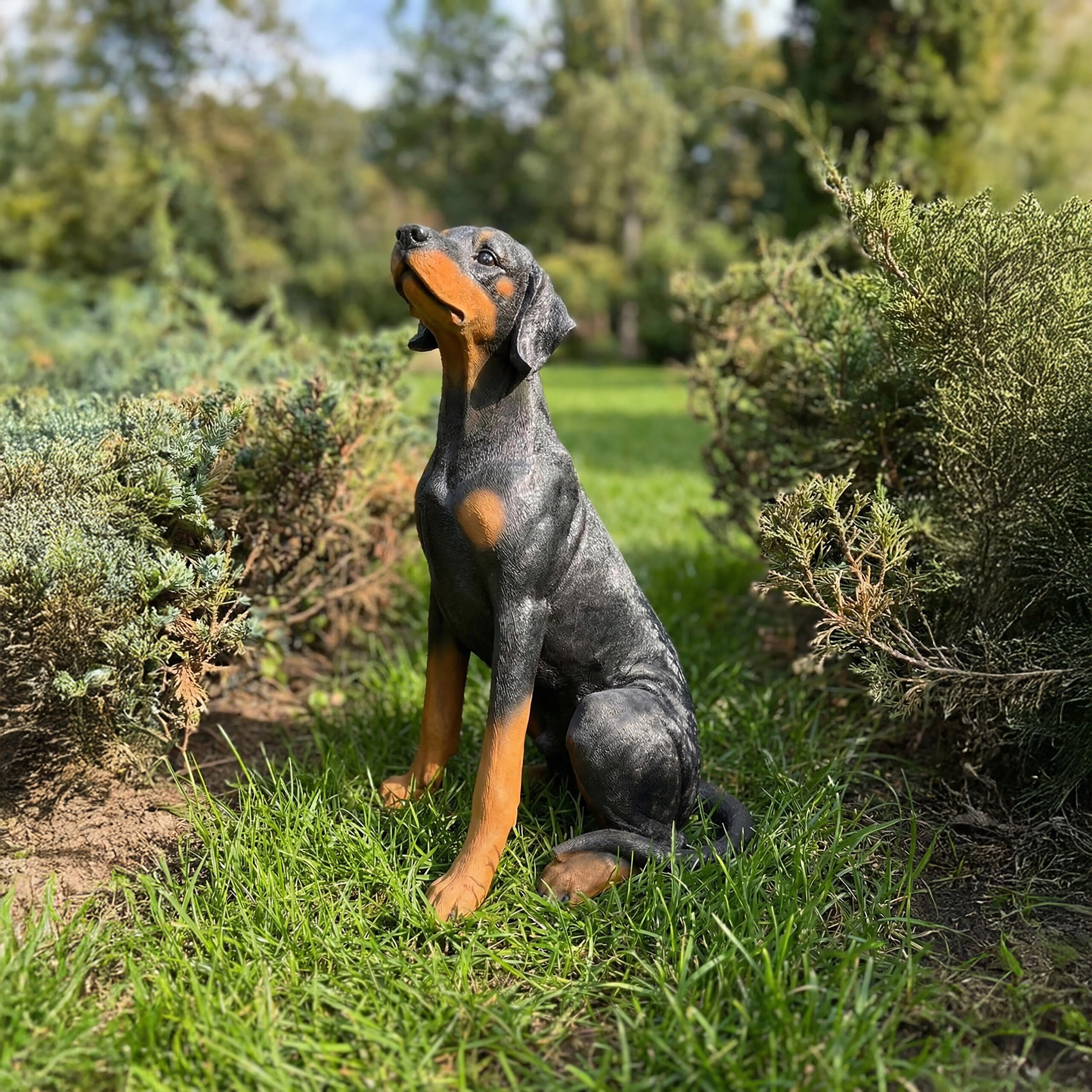 Eine schwarz-braune Dobermann-Hundefigur sitzt im Gras zwischen Sträuchern.