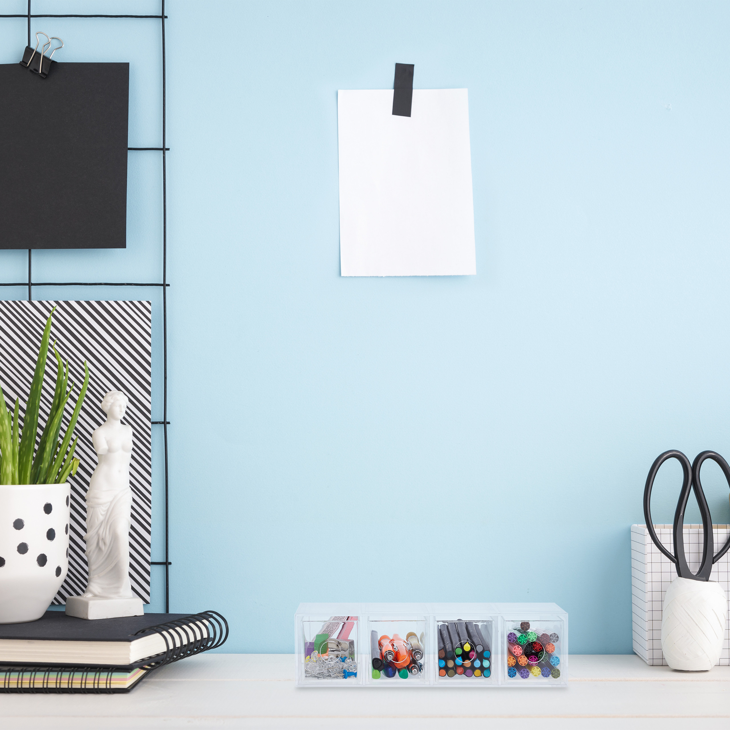 Ein ordentlicher Schreibtisch mit hellblauer Wand, Notizblättern, Stiftebox, Schere und kleinen Deko-Objekten.