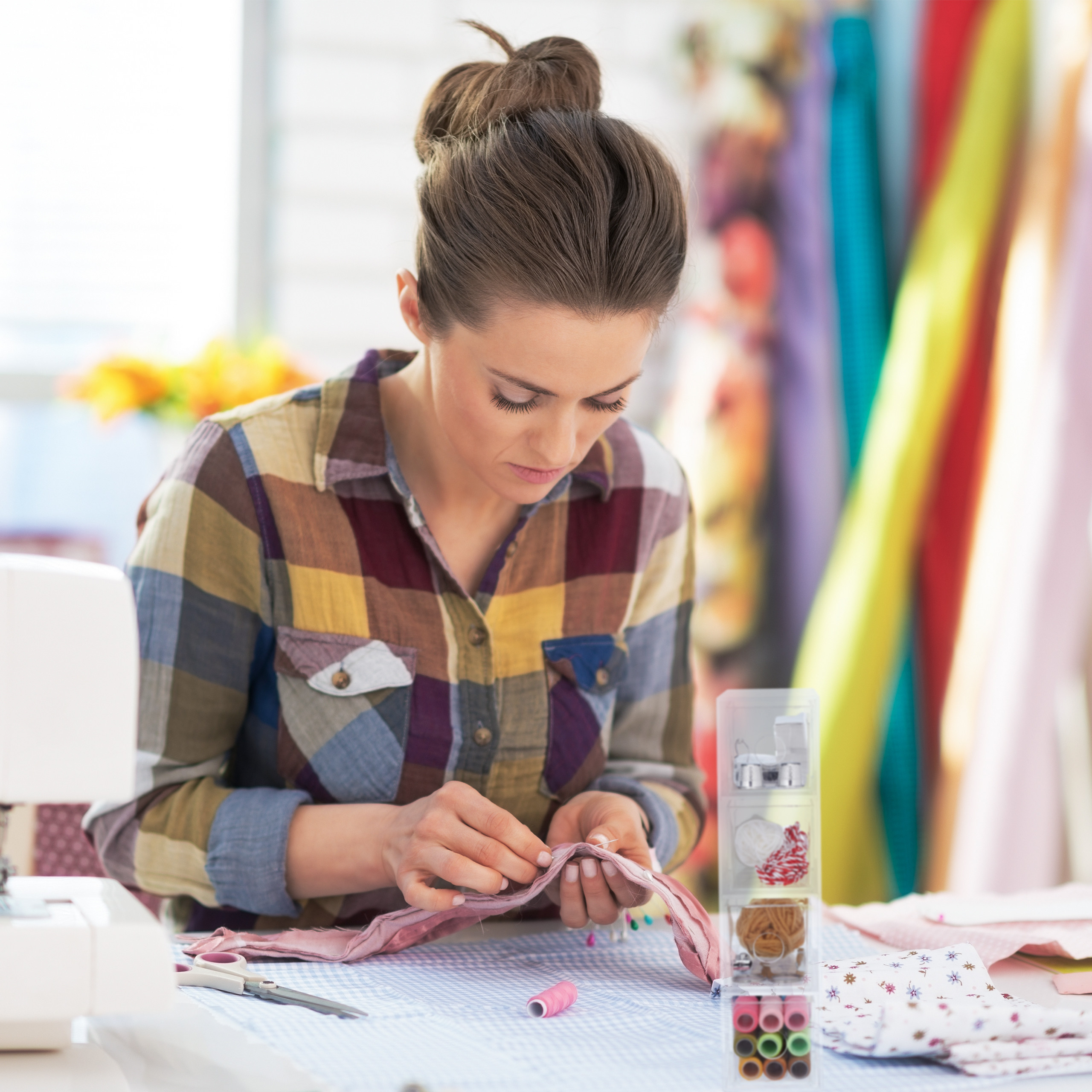 Eine Frau im karierten Hemd näht in einem Atelier mit bunten Stoffen im Hintergrund.