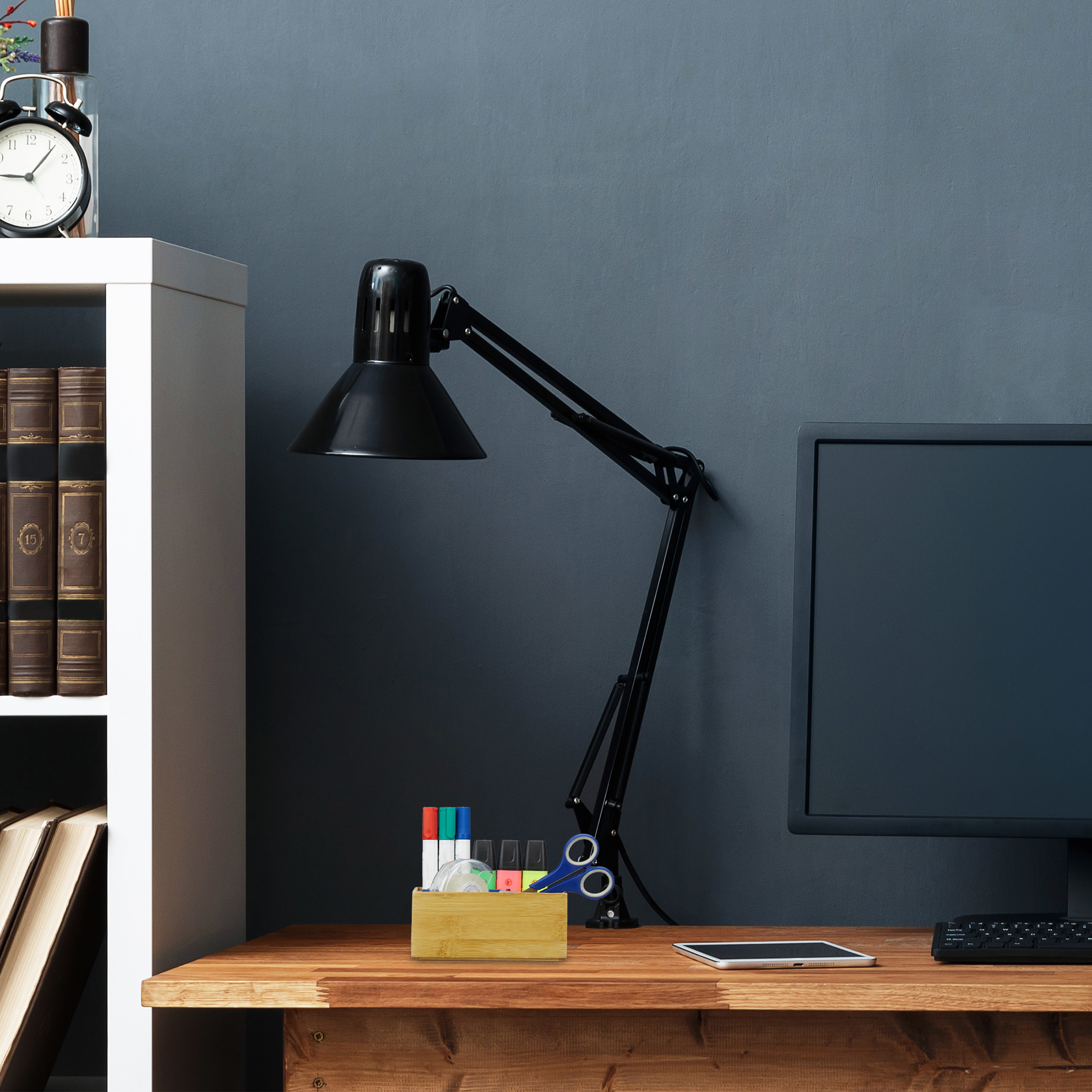 Ein Schreibtisch mit schwarzer Lampe, Monitor, Stifteköcher, Büchern und einer dunkelgrauen Wand im Hintergrund.